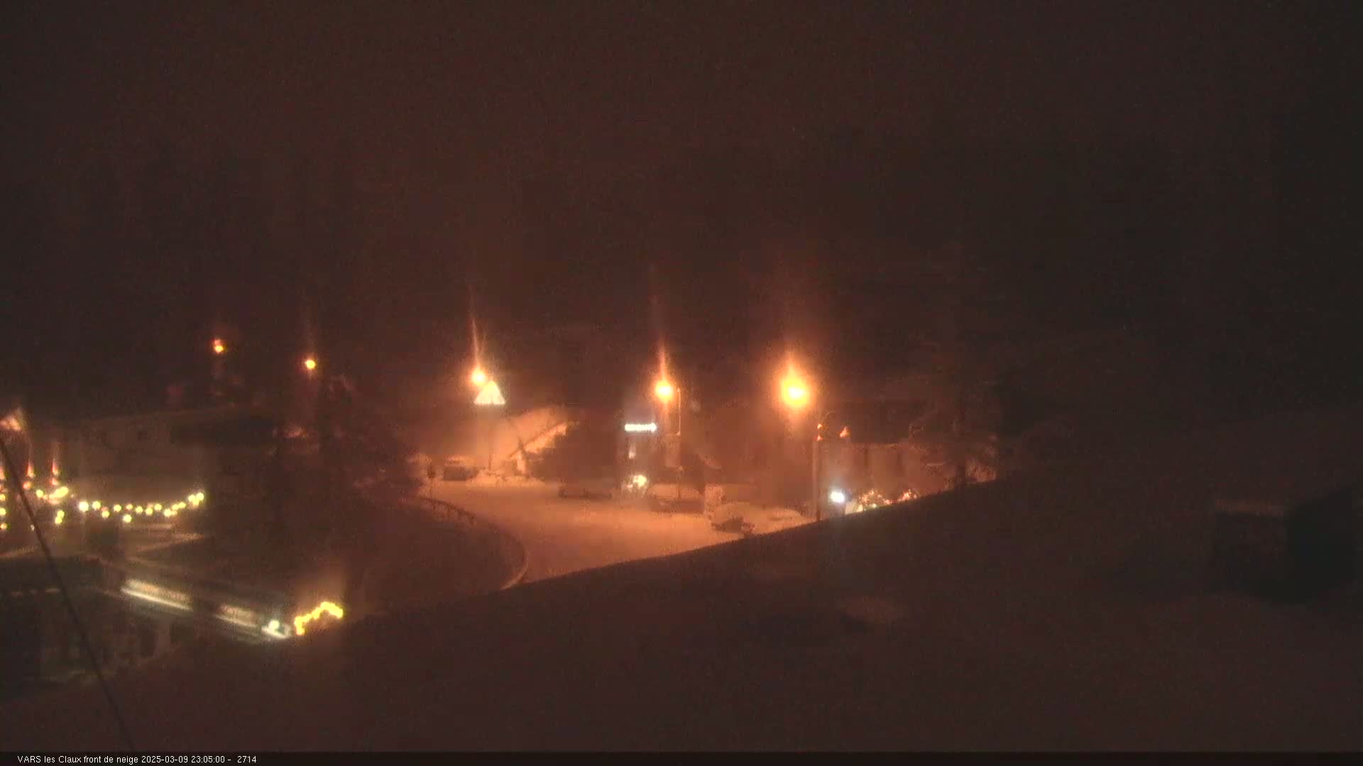 A snow-covered parking lot at night is dimly lit by streetlights in hazy conditions.