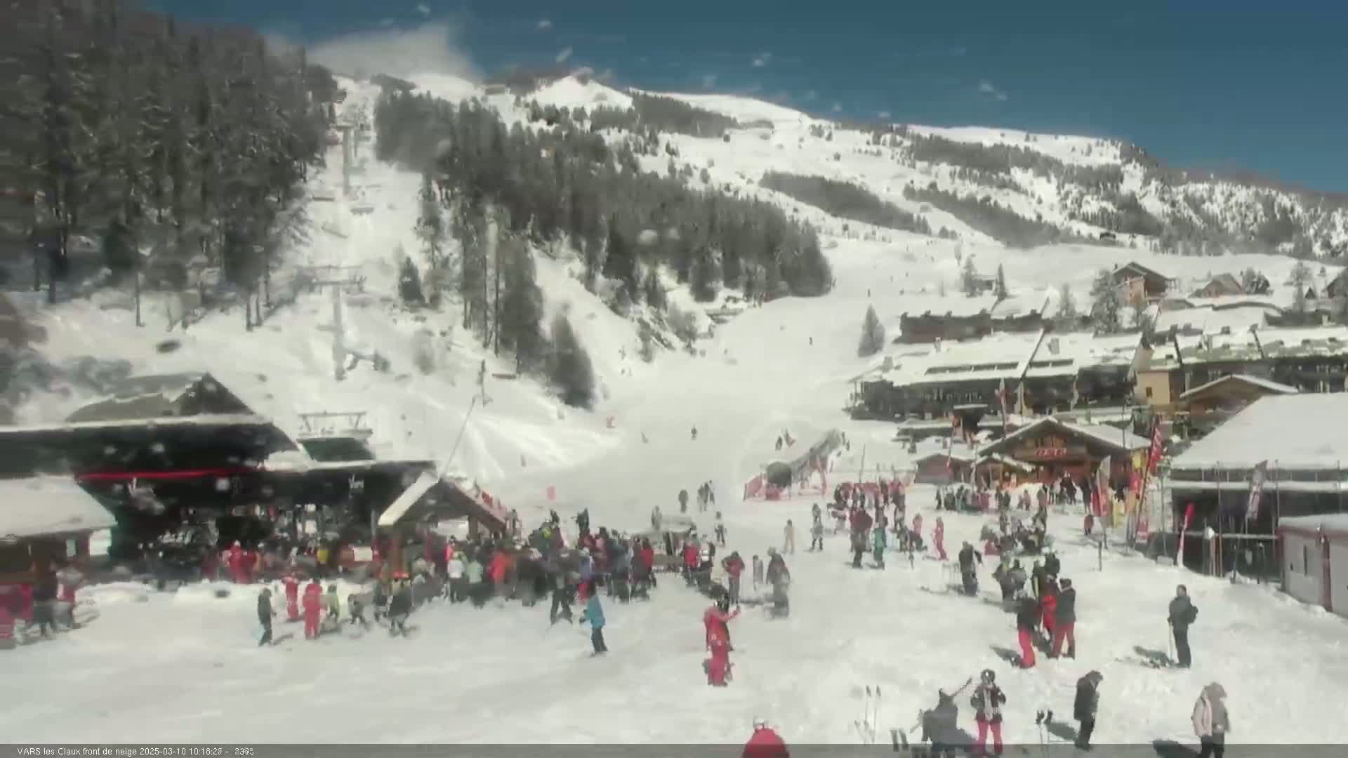 A snowy ski resort with many people skiing and gathered near buildings on a sunny day.