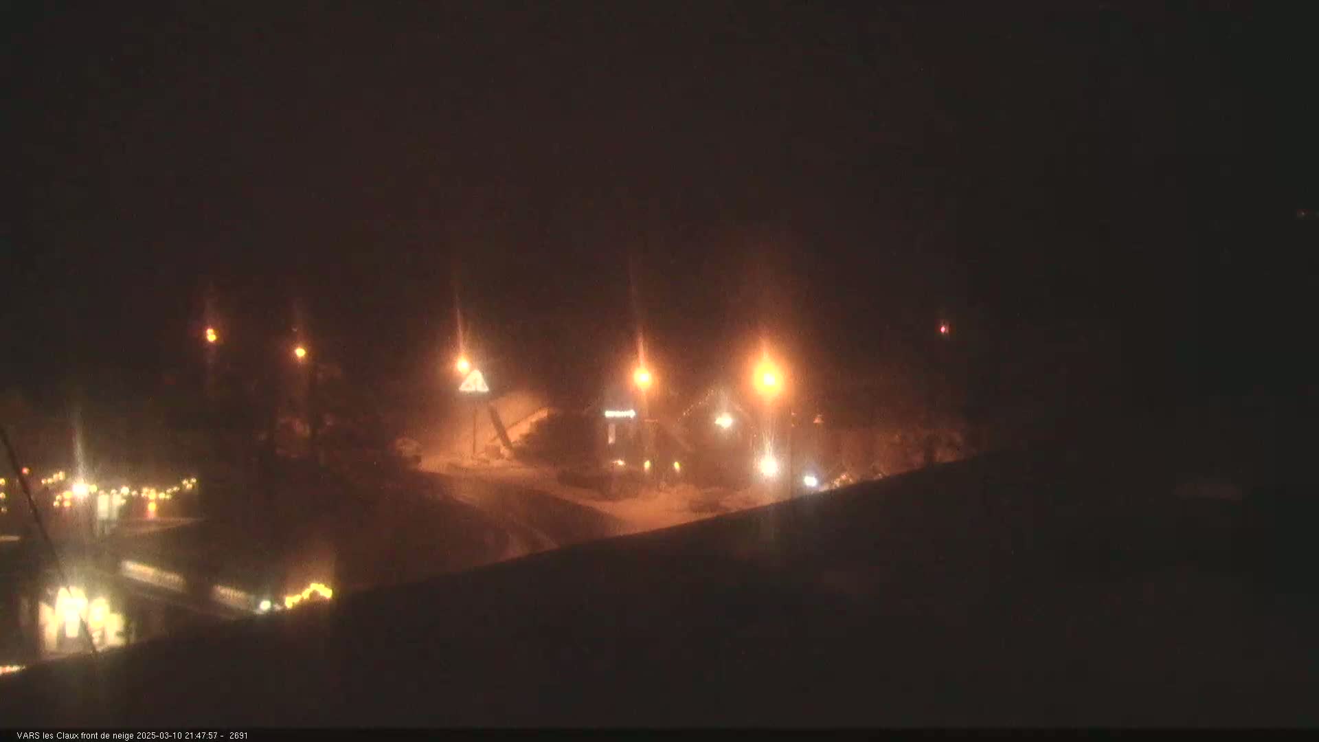 A nighttime, snow-covered scene is illuminated by several streetlights, showing a road with vehicles and buildings in the distance.