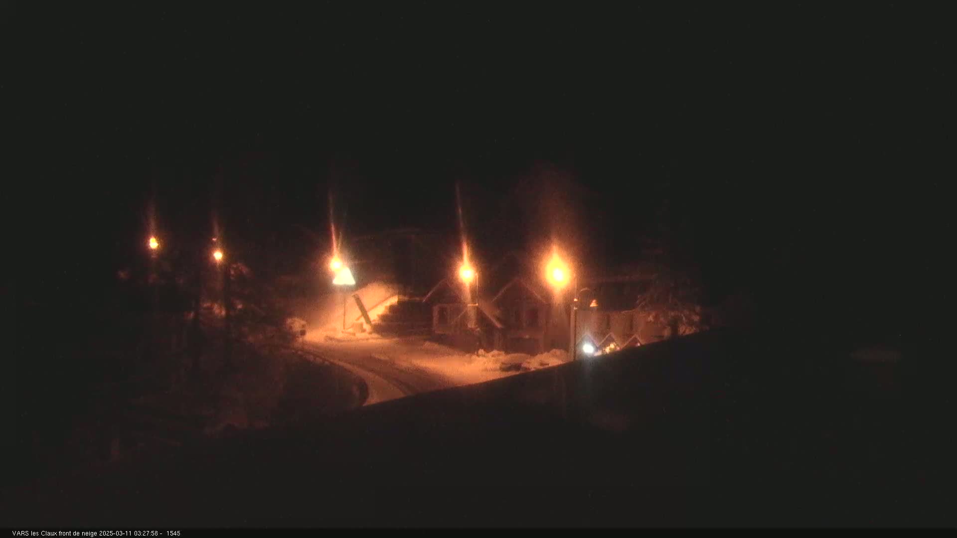 A nighttime, snowy scene shows several illuminated buildings along a curving road.