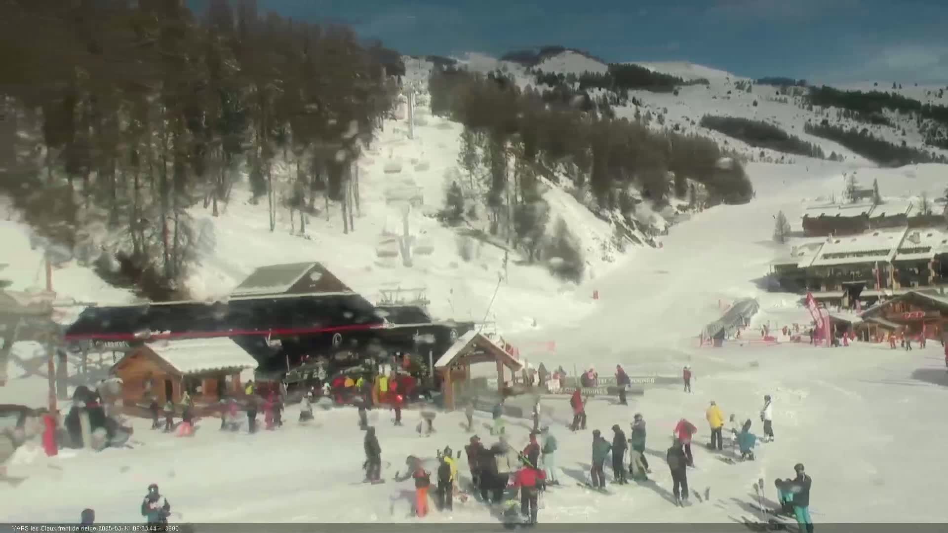 A snowy ski resort on a sunny day shows skiers and snowboarders at the base of a slope near several buildings.