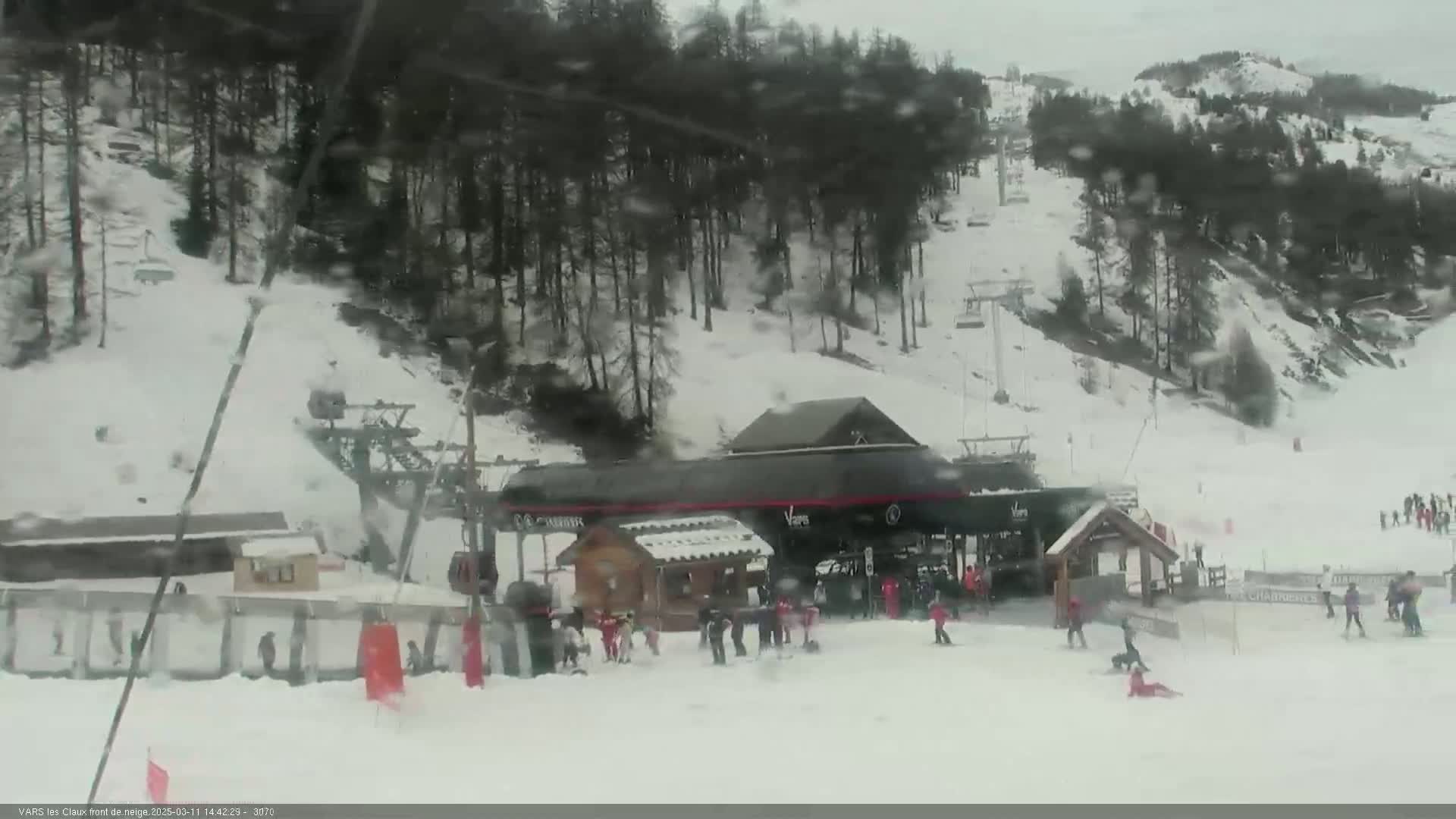 A snowy ski resort scene shows a ski lift base area with people skiing and snowboarding in light snow.