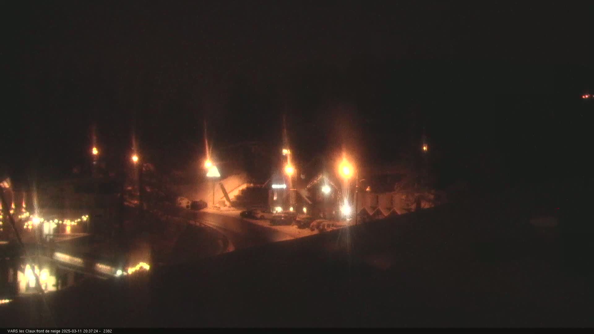 A nighttime scene showing a snow-covered area with several illuminated buildings and streetlights.