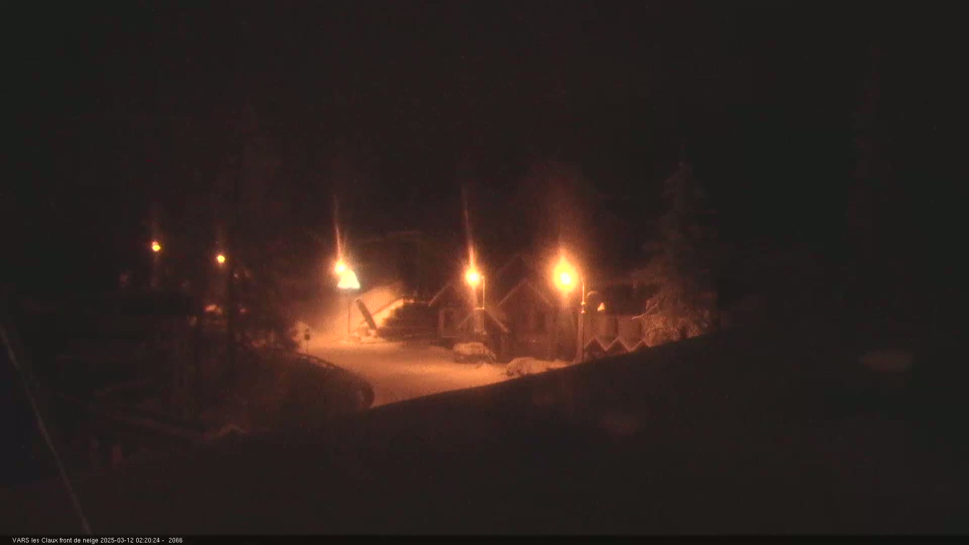 A nighttime scene of snow-covered ground illuminated by several lampposts near a cluster of buildings.