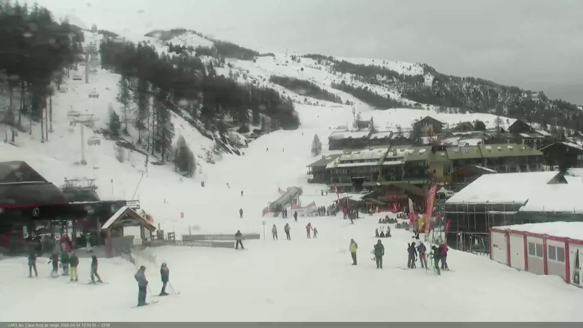 A snowy ski resort with several skiers and snowboarders on the slopes under an overcast sky.