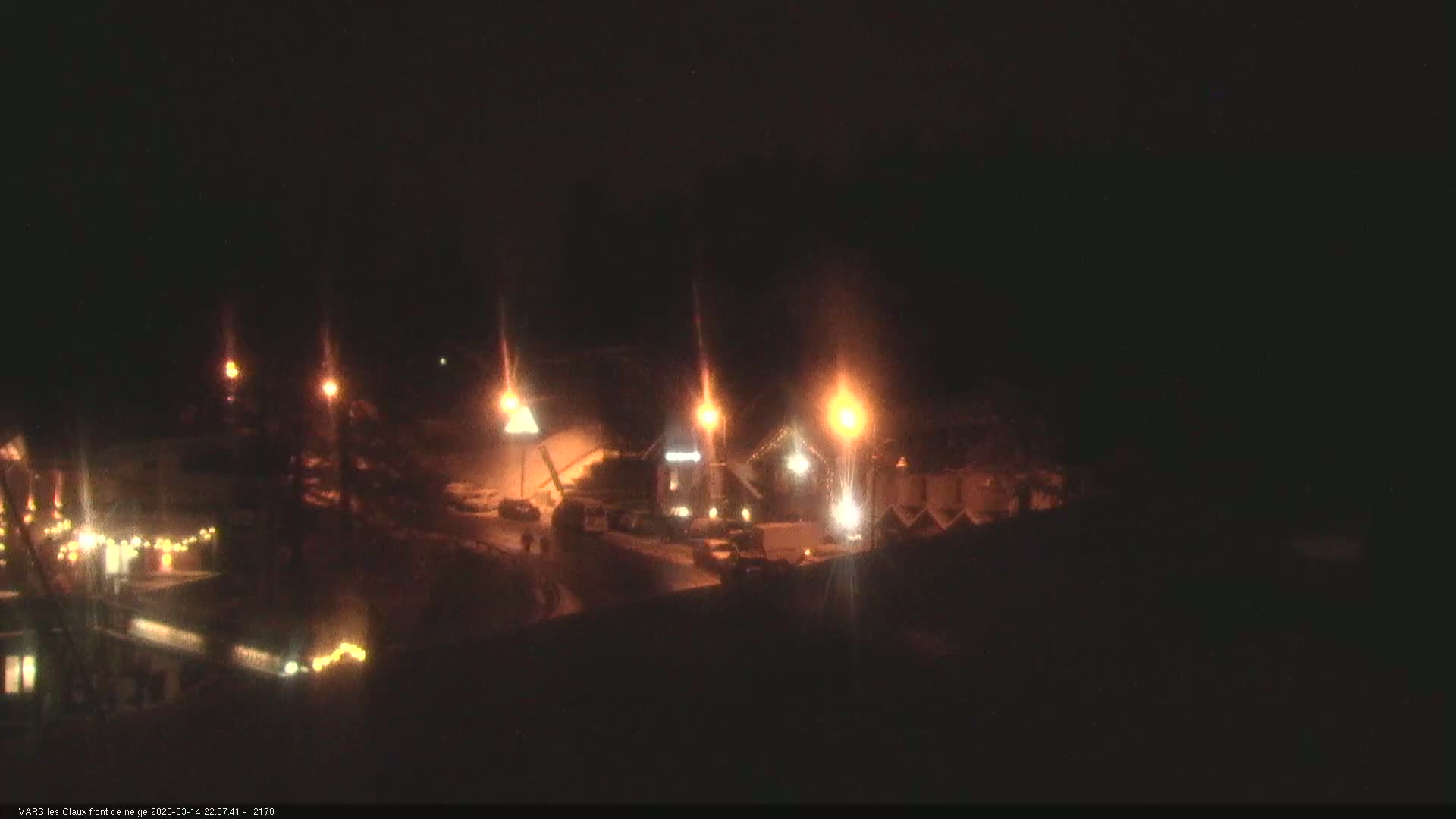 A nighttime scene shows a snow-covered area with several illuminated buildings and streetlights.