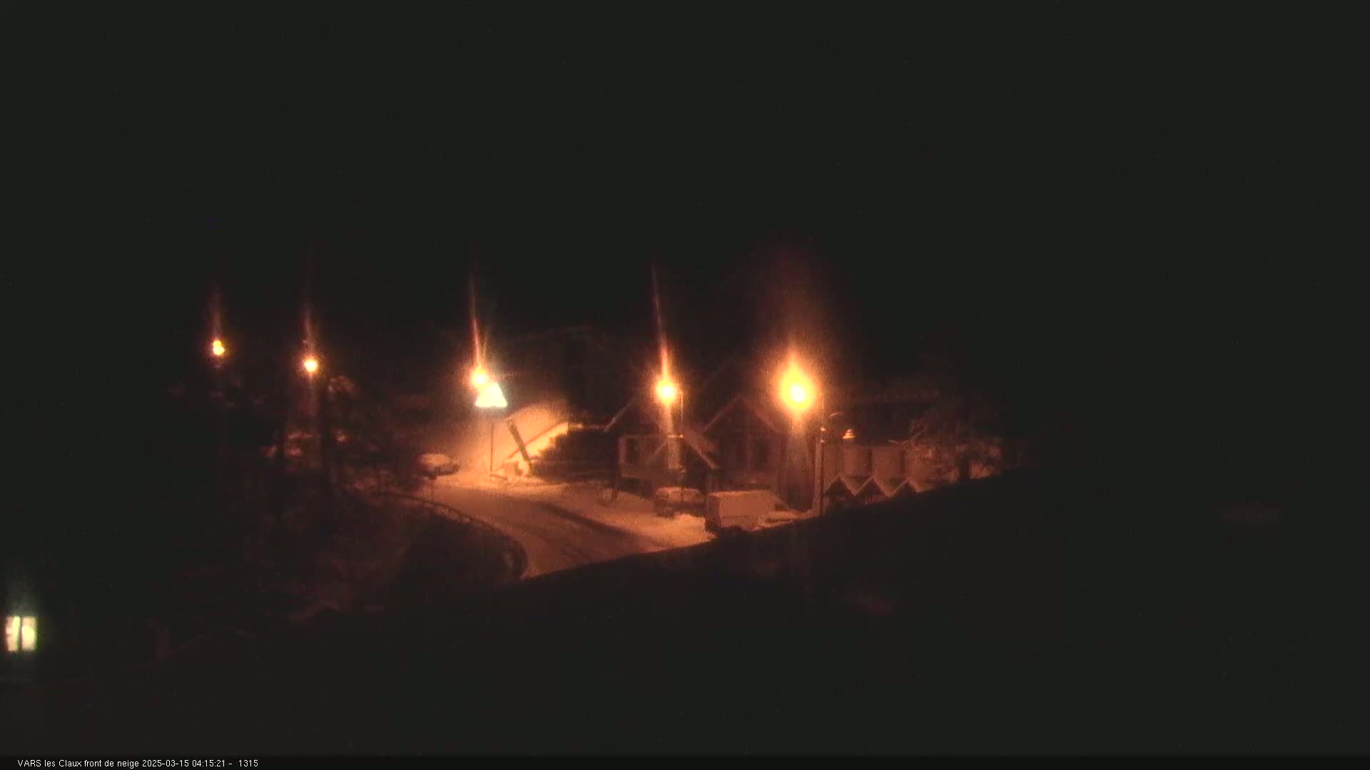 At night, under snowy conditions, illuminated buildings and a road are visible, with several streetlights casting a warm glow.