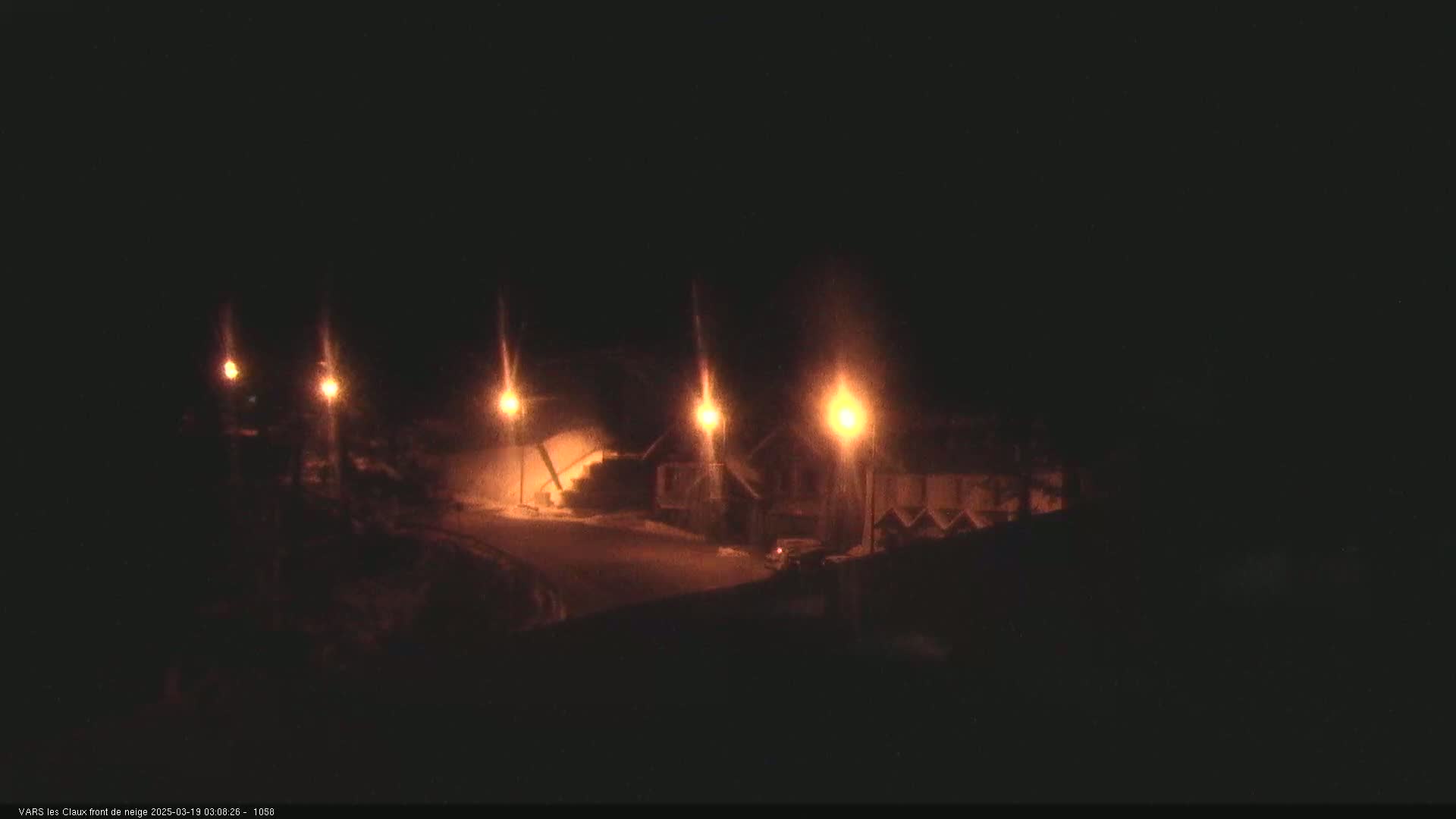 A nighttime, snow-covered scene shows several illuminated streetlights lining a road in front of buildings.