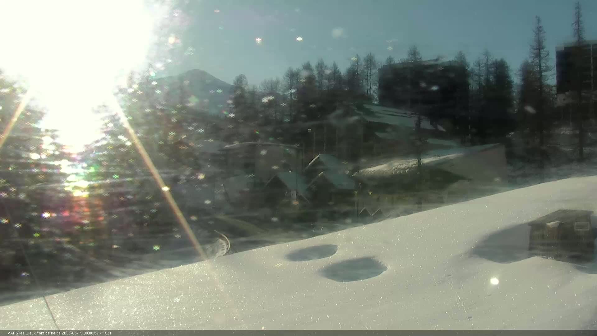 A snow-covered landscape with buildings and trees in the background, viewed on a bright, sunny day with some glare from the sun.