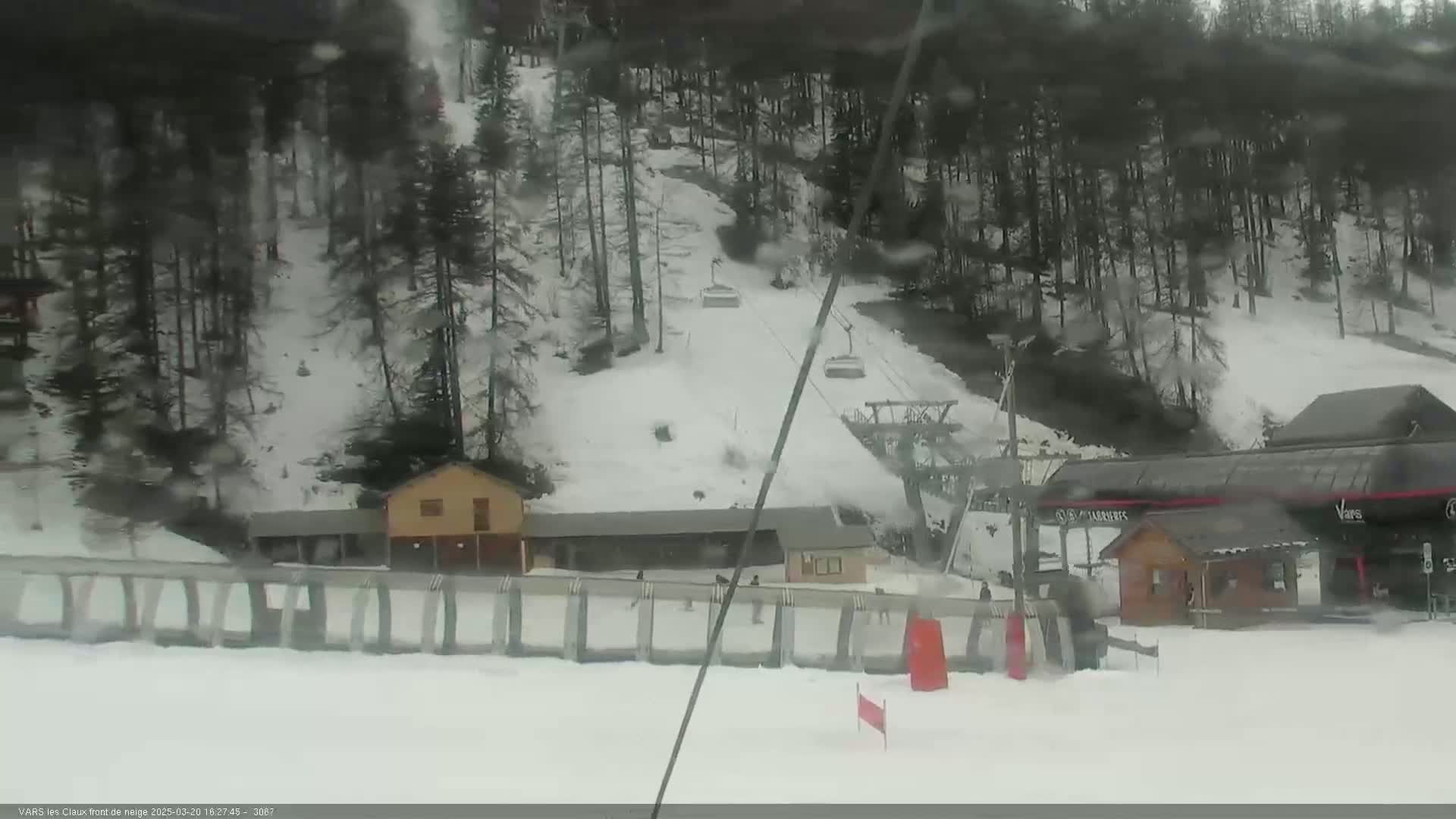 A snow-covered ski slope with a chairlift and several small buildings is visible through a slightly blurry window, in overcast weather.