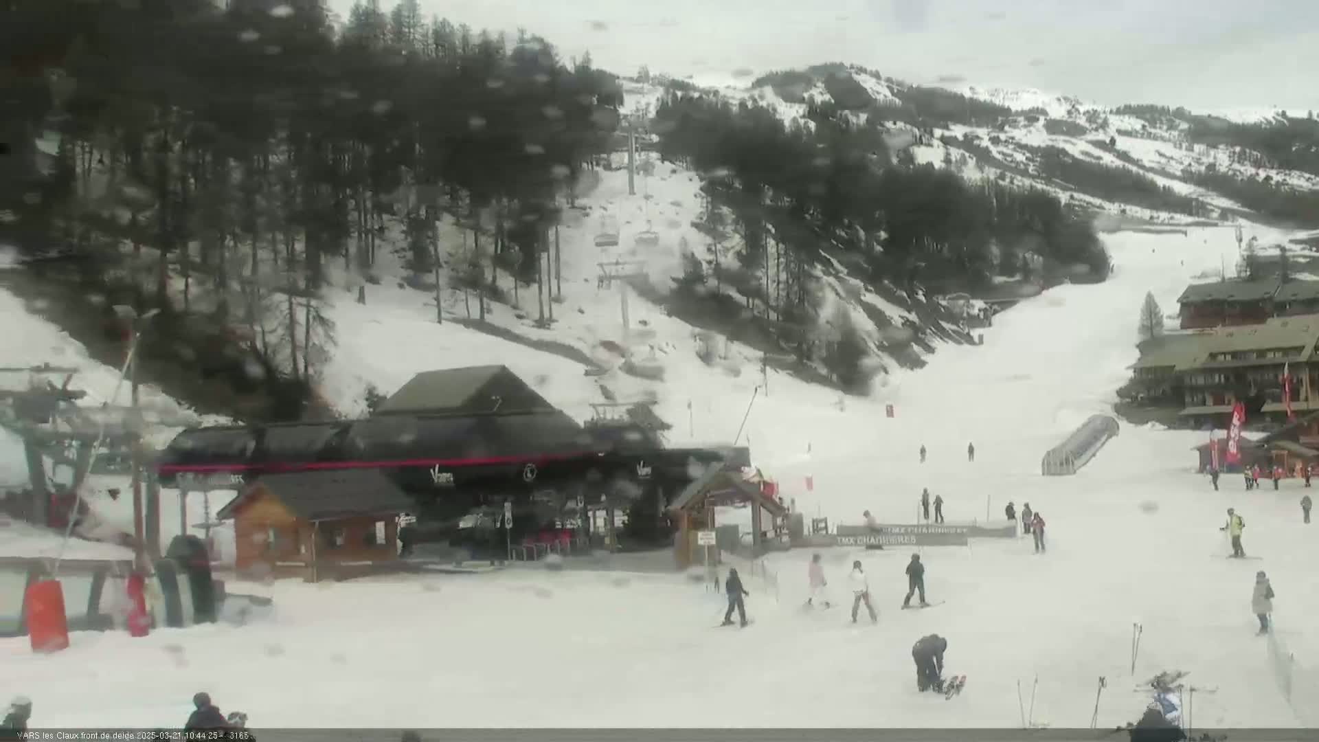 A snow-covered ski slope with several skiers and snowboarders, a ski lift, and buildings, under a cloudy, snowy sky.