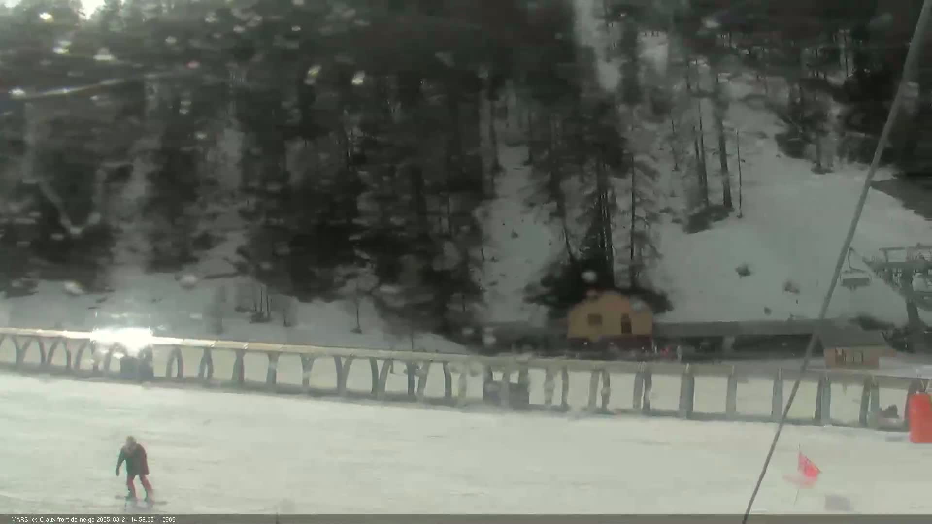 A snowy, lightly falling snow scene shows a person snowboarding down a slope near a fence and trees.