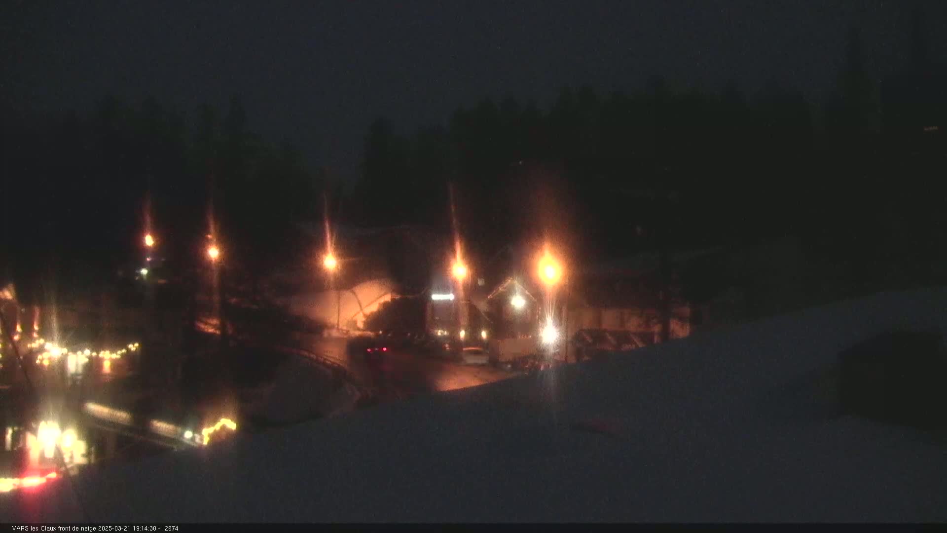 A nighttime scene of a snow-covered road with vehicles and streetlights illuminated against a dark, wooded backdrop.