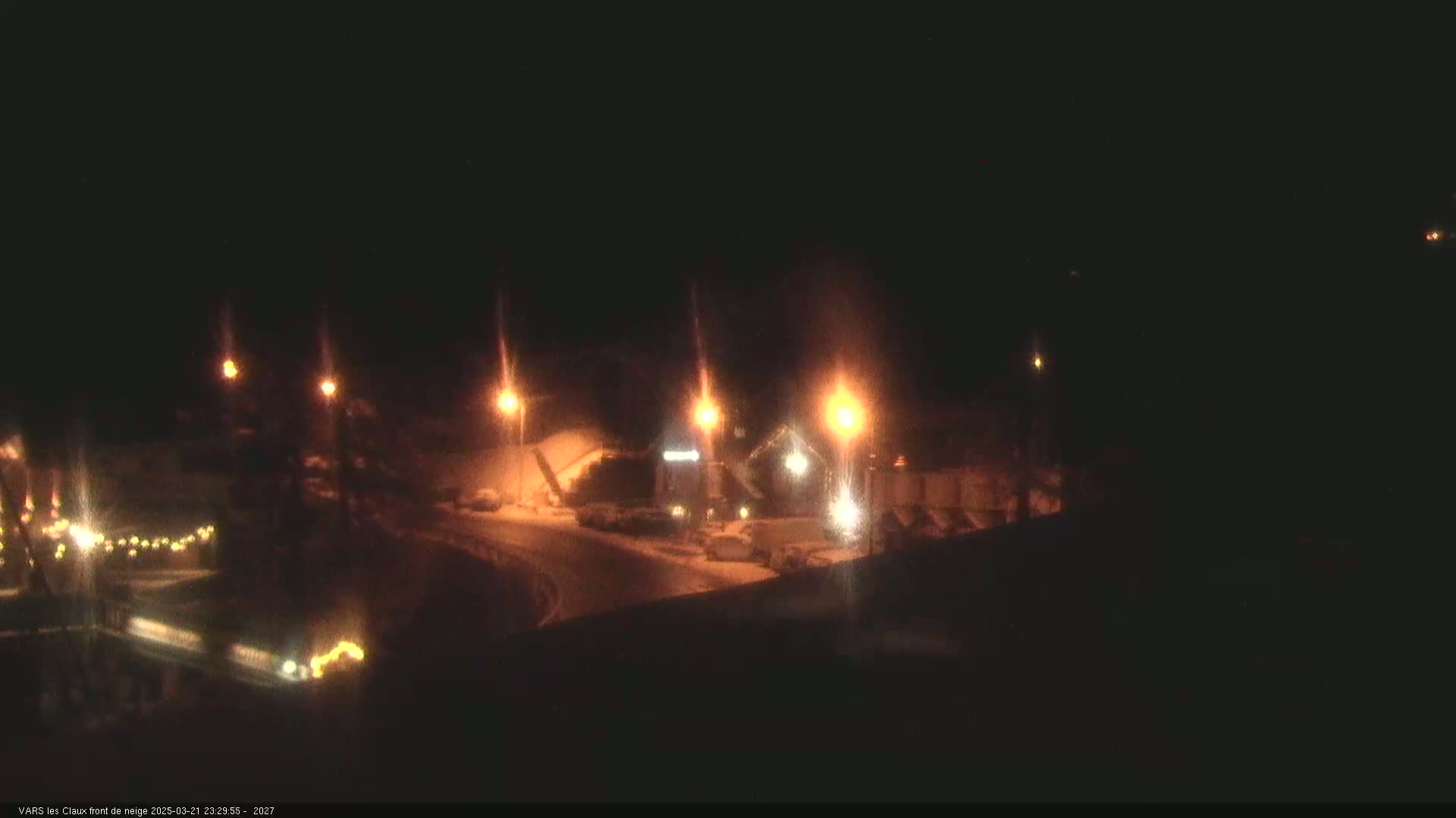 A nighttime, snowy scene shows a roadway curving around a building complex illuminated by streetlights.
