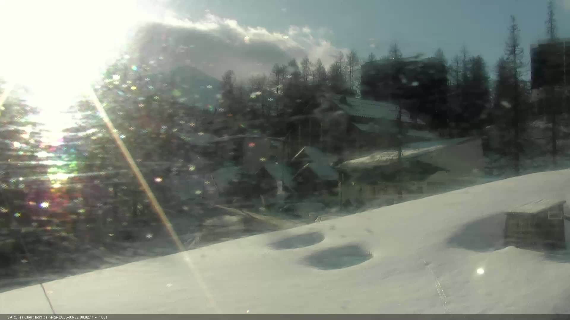 A sunny, snowy landscape shows a village nestled among evergreen trees, with some buildings partially obscured by bright sunlight.