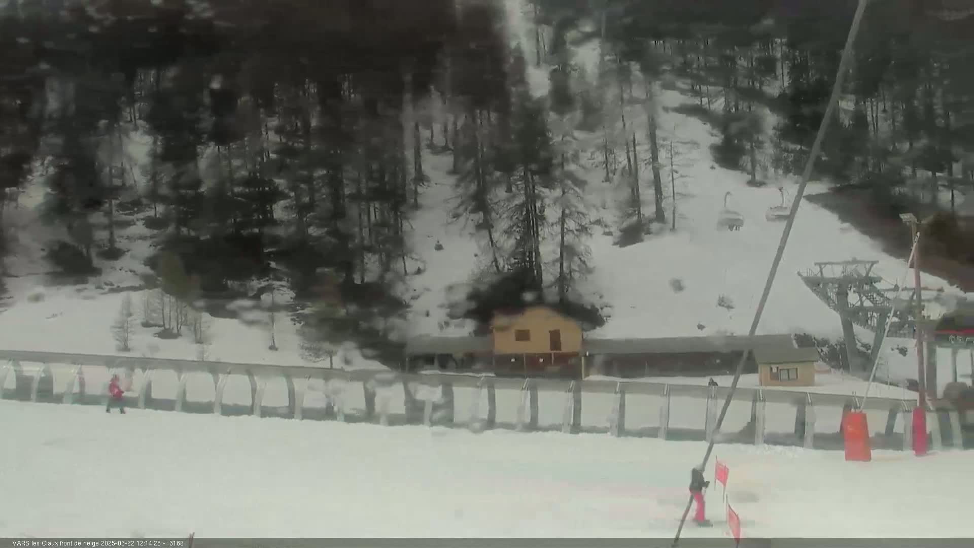 A snowy ski slope with a small building, a ski lift, and a few people skiing on an overcast day.