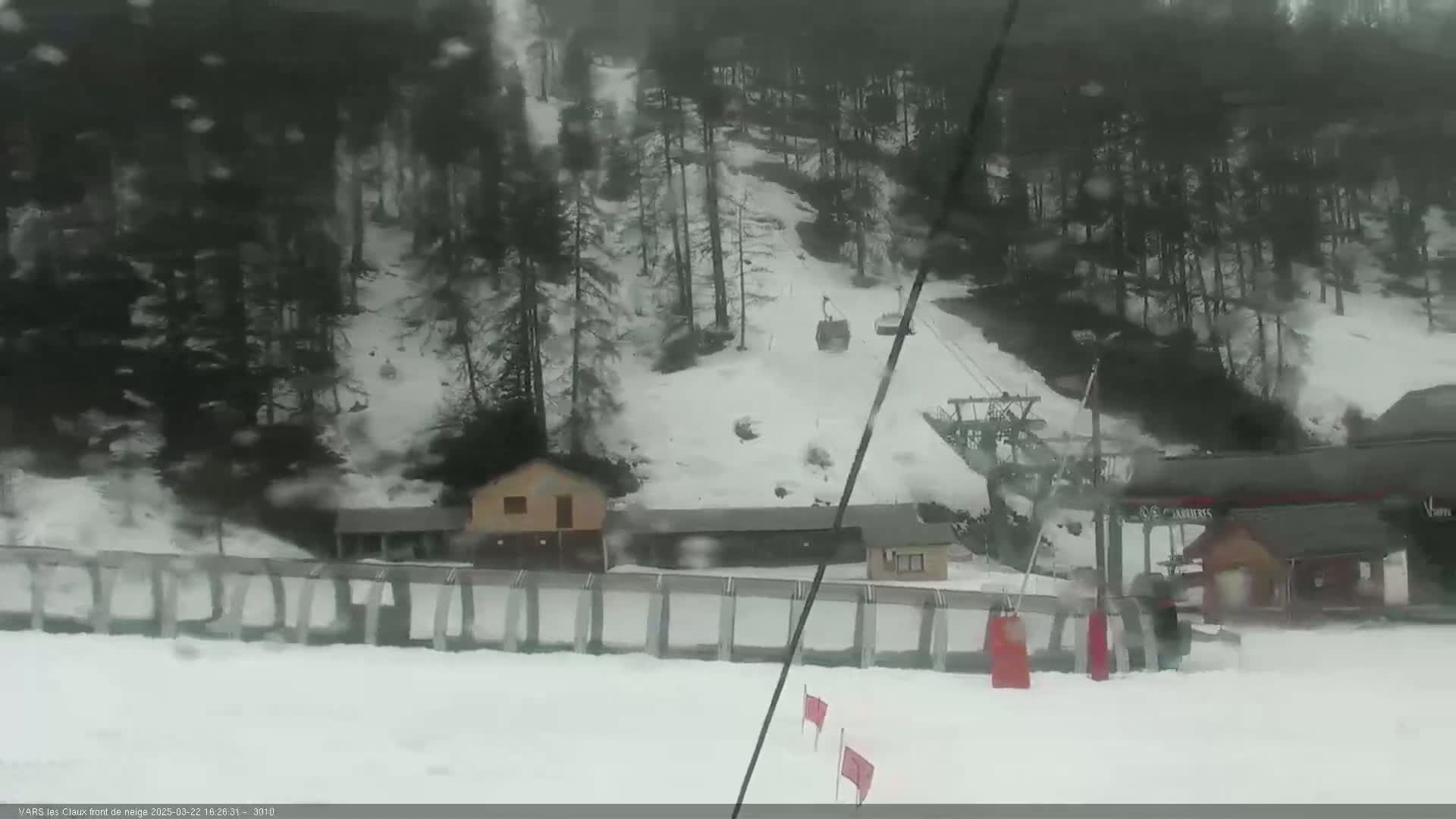 A snowy ski slope with a chairlift and small buildings is seen through a snow-filled atmosphere.