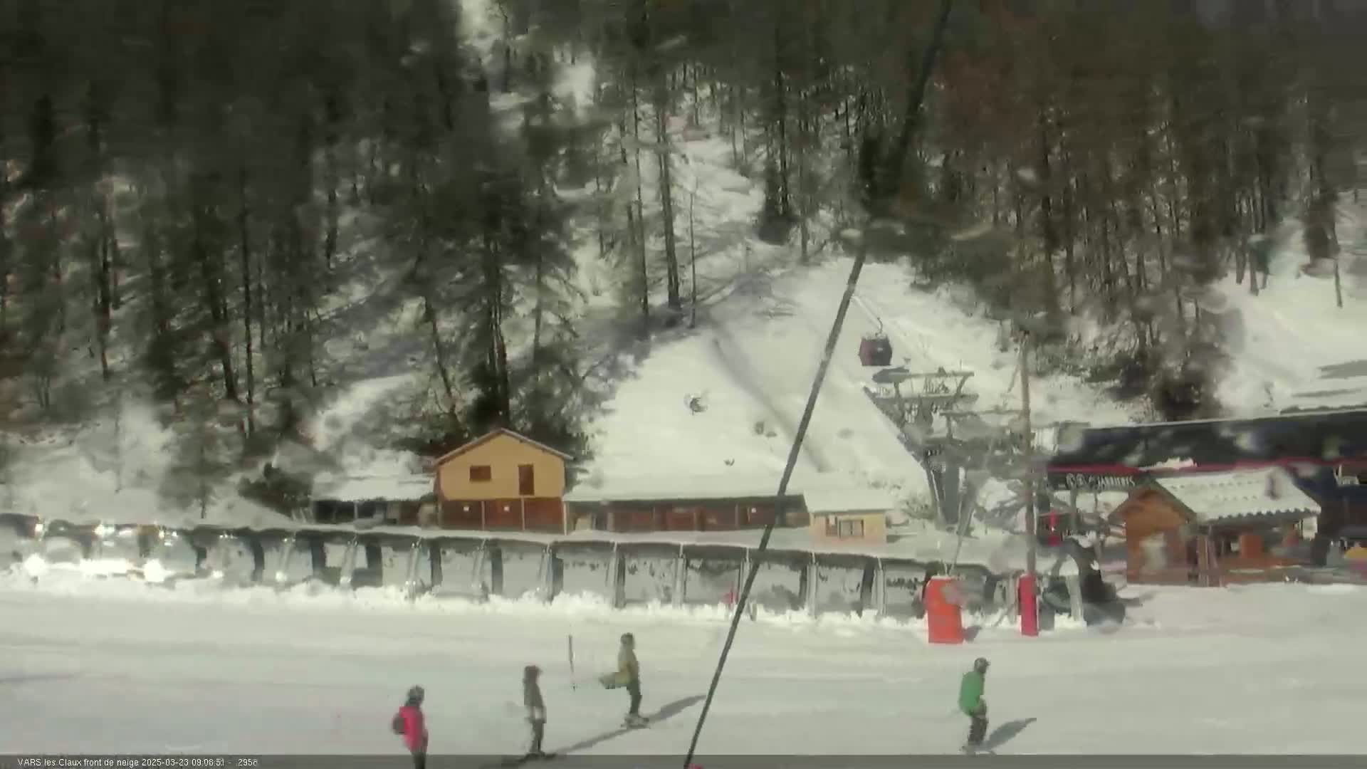 A snowy ski slope with several skiers, a ski lift, and small buildings nestled among snow-covered trees under a bright, sunny sky.