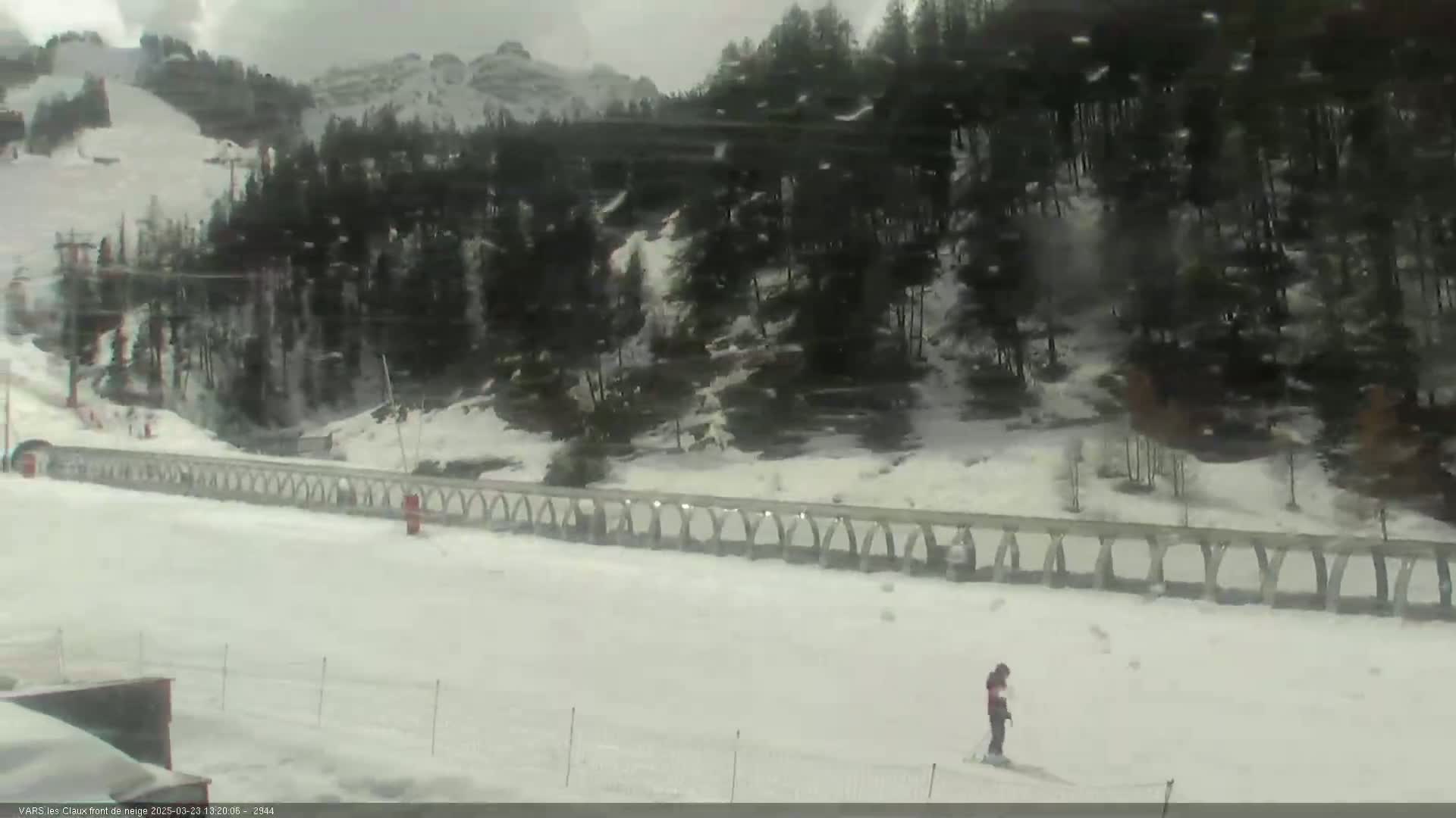 A snowy ski slope with a person skiing in the foreground, a low stone wall behind them, and a snow-covered forest and mountains in the background under an overcast sky.