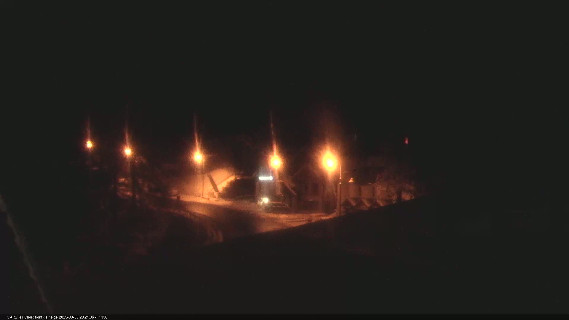 A nighttime scene shows a snow-covered road illuminated by several streetlights, with a vehicle visible in the distance.