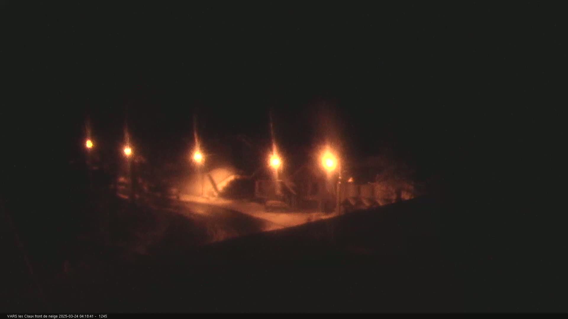 At night, under snowy conditions, several illuminated structures are visible, possibly buildings or lampposts.