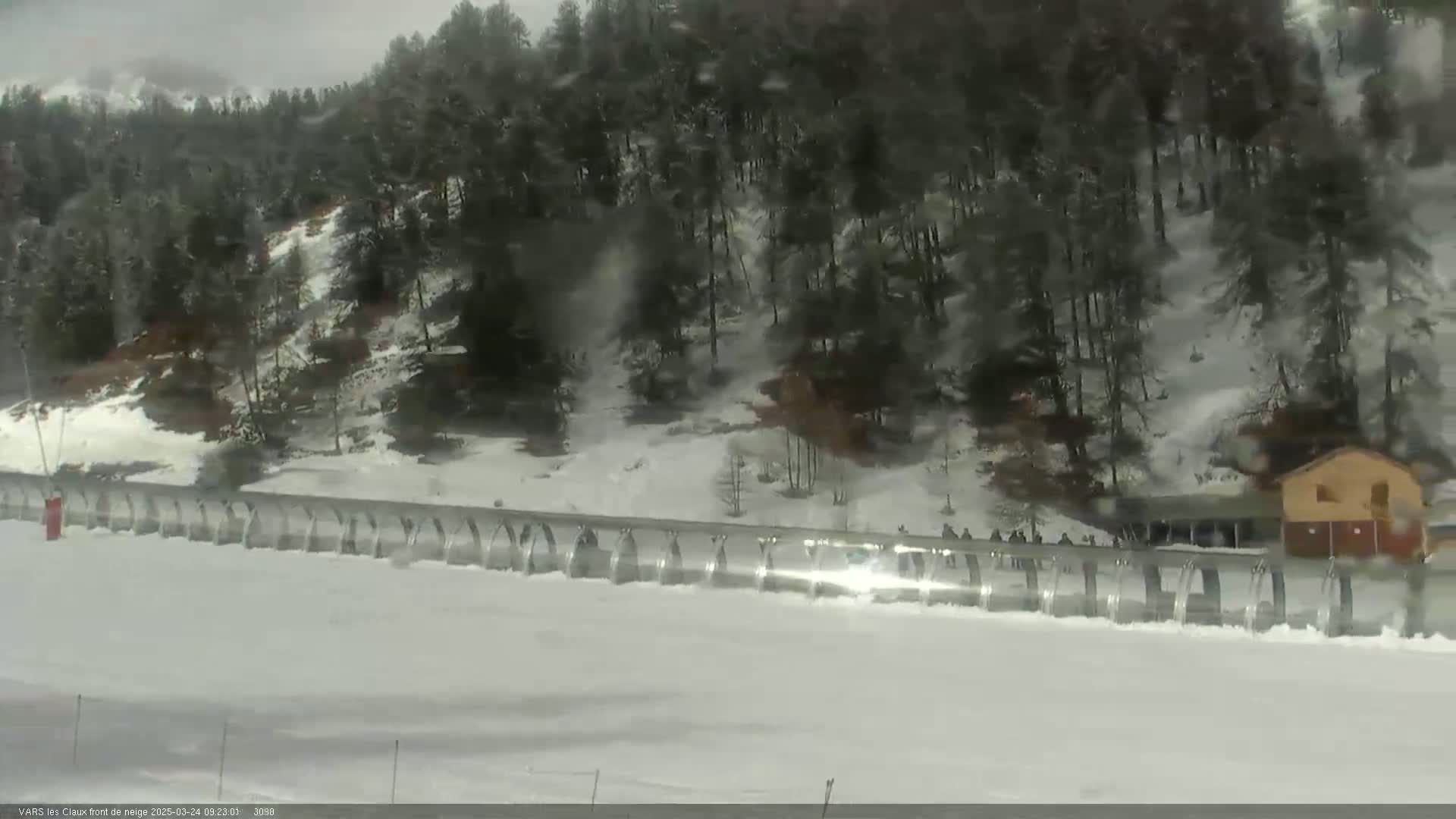 A snowy landscape with a long, curved barrier stretches across a flat expanse of snow, behind which stand snow-covered evergreen trees and a small building, under overcast skies.