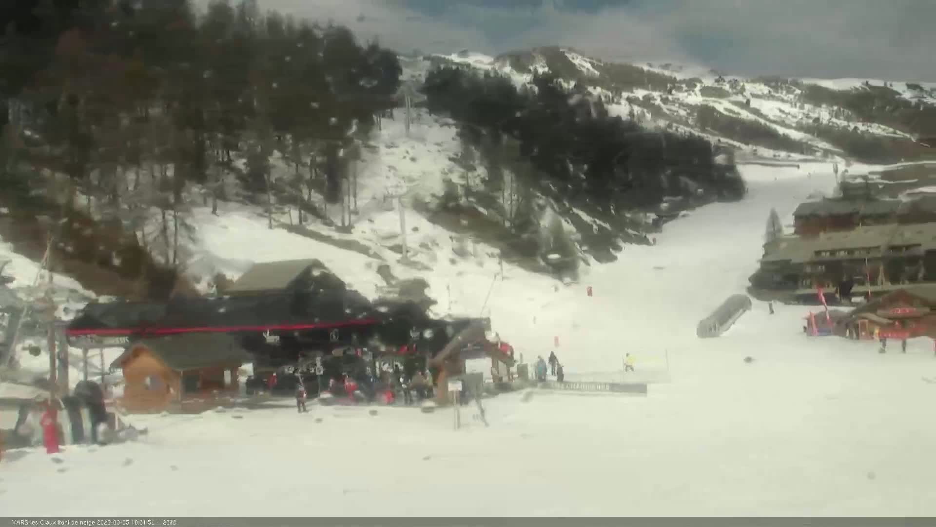 A snowy ski slope with a lift station, several skiers, and buildings in the background under a partly cloudy sky.