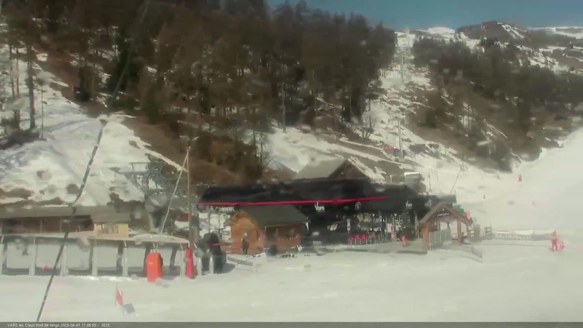 A ski resort on a sunny day shows a snow-covered slope with a ski lift and a small building near the bottom of the hill, and a few skiers are visible.