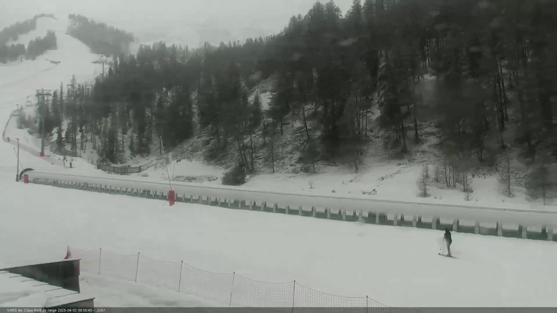 A snowy ski slope with a long row of covered structures and a single skier visible in the lower right, under overcast skies.