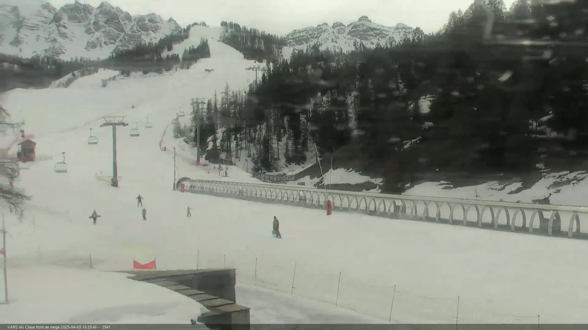A snowy ski slope with several people skiing and snowboarding, under an overcast sky, is bordered by trees and mountains in the background.