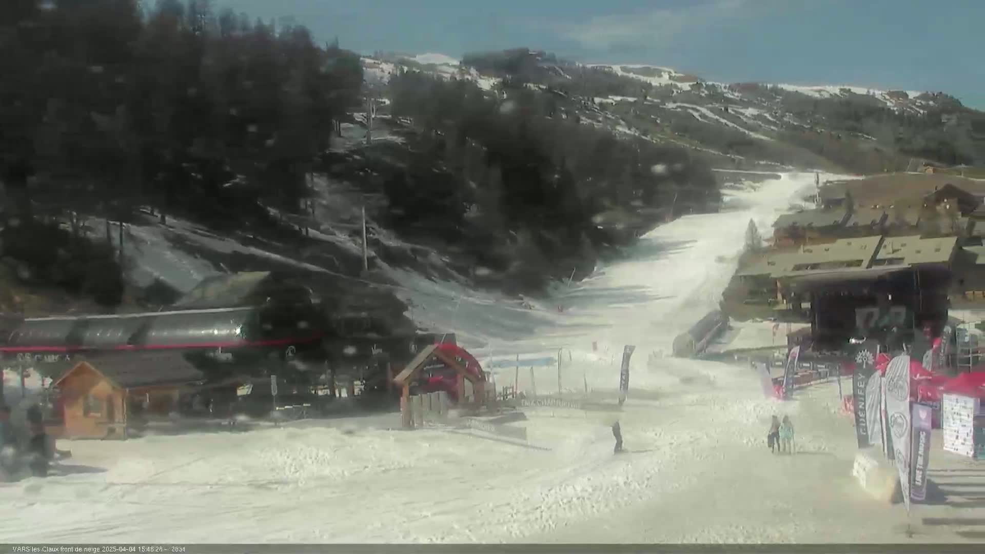 A snow-covered ski slope with several buildings, a ski lift, and a few people are visible on a partly sunny day.