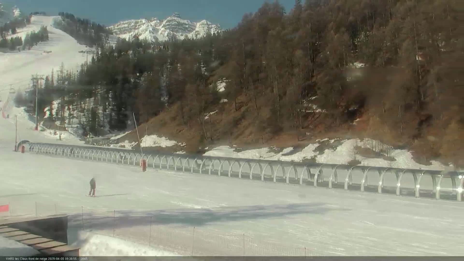 A snowy, sunny ski slope with a long row of arched structures and a person skiing in the foreground, backed by a mountain range and a forest.