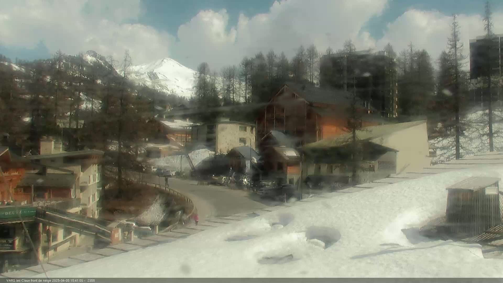 A partly cloudy day reveals a snow-covered mountain village nestled amongst evergreen trees and a snow-capped mountain in the distance.