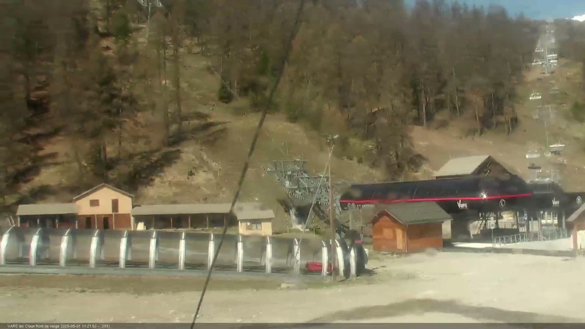 A ski lift base area with several buildings, ski racks, and a chairlift ascending a hillside covered in mostly bare, brown trees under a sunny sky.