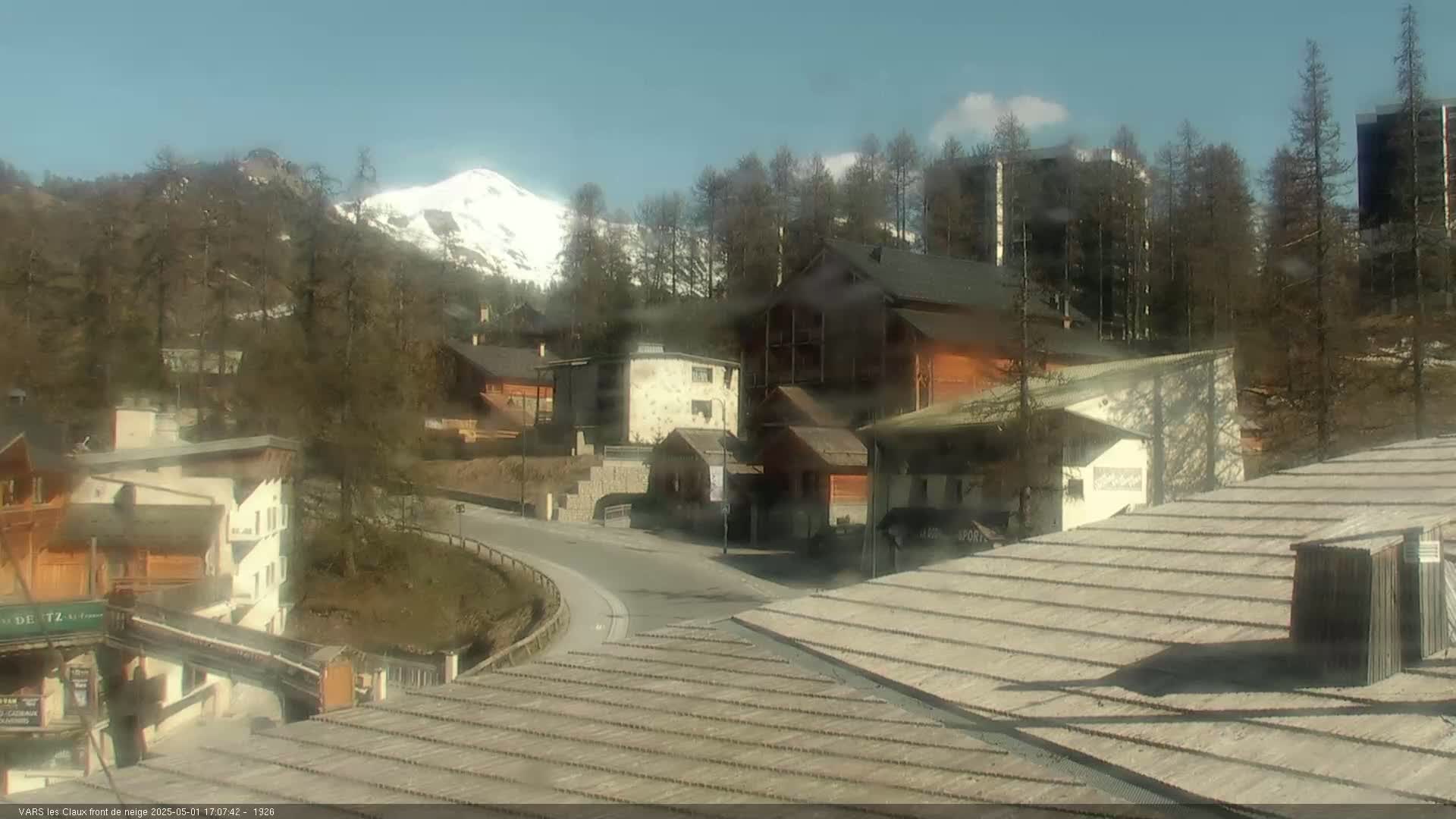 A mountain village nestled among trees, with a snow-capped mountain in the background under a clear, sunny sky.