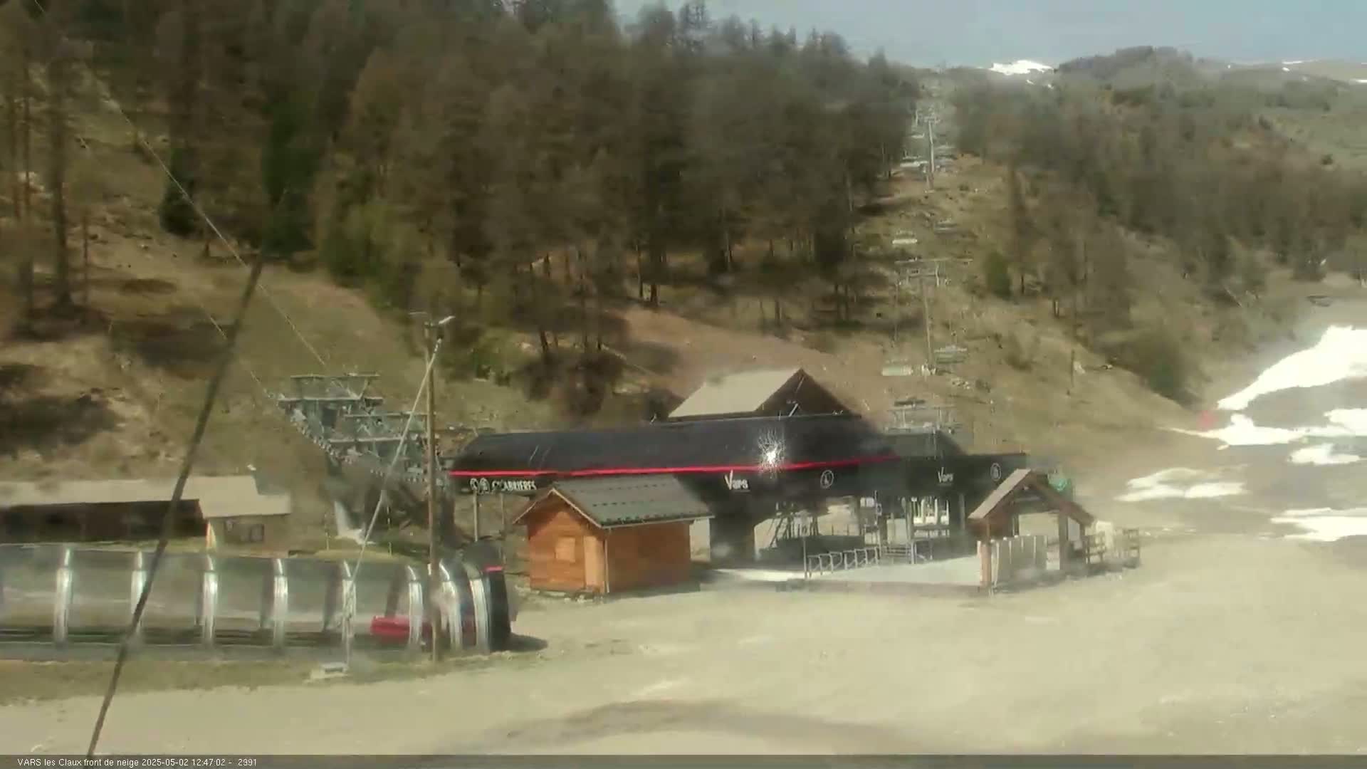 A ski lift base station with a small building and a long, covered conveyor belt sits on a mostly barren hillside under a sunny sky.