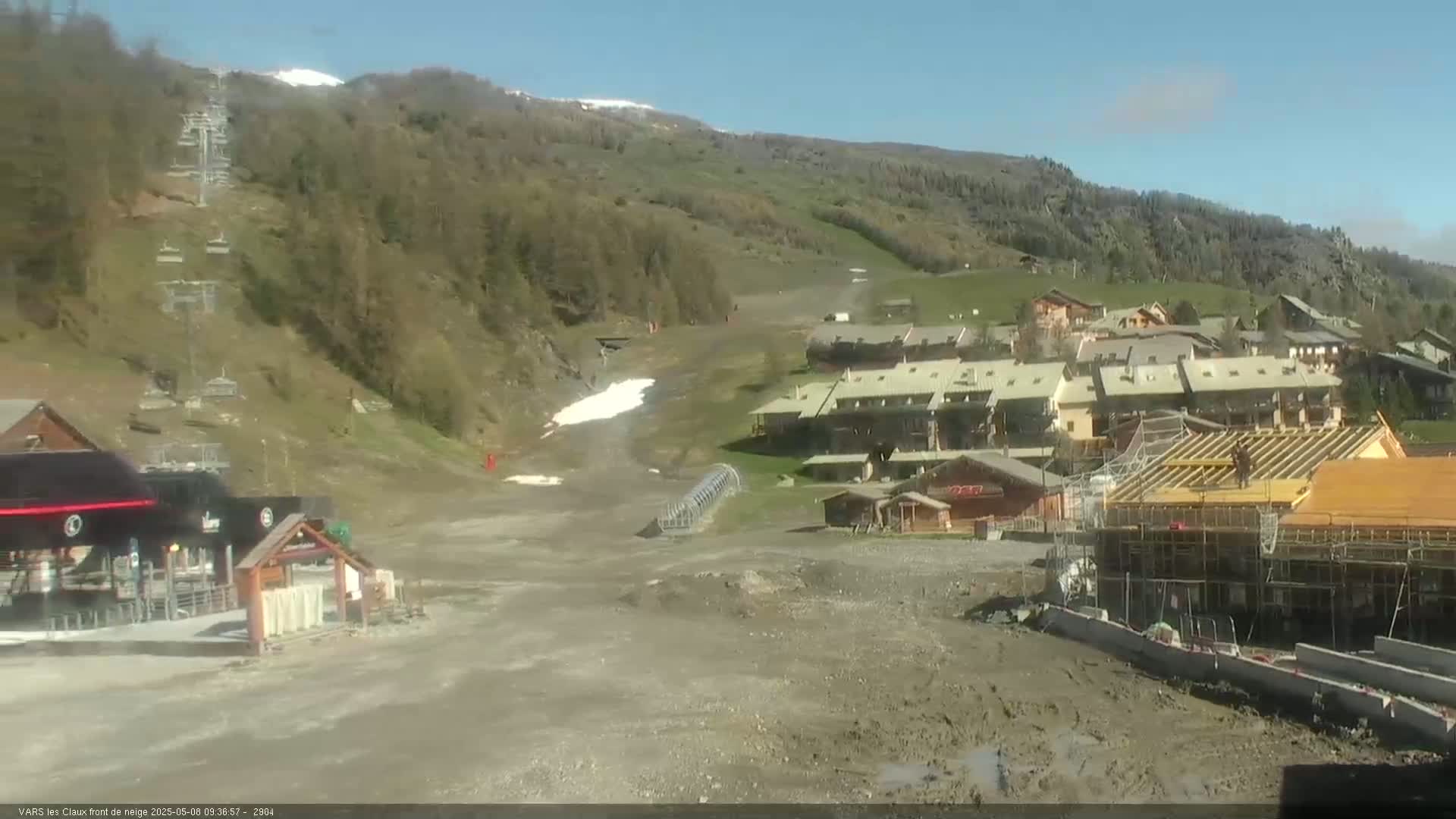 A mostly clear day reveals a ski resort with a chairlift, buildings, and a construction site, situated on a hillside with patches of remaining snow.