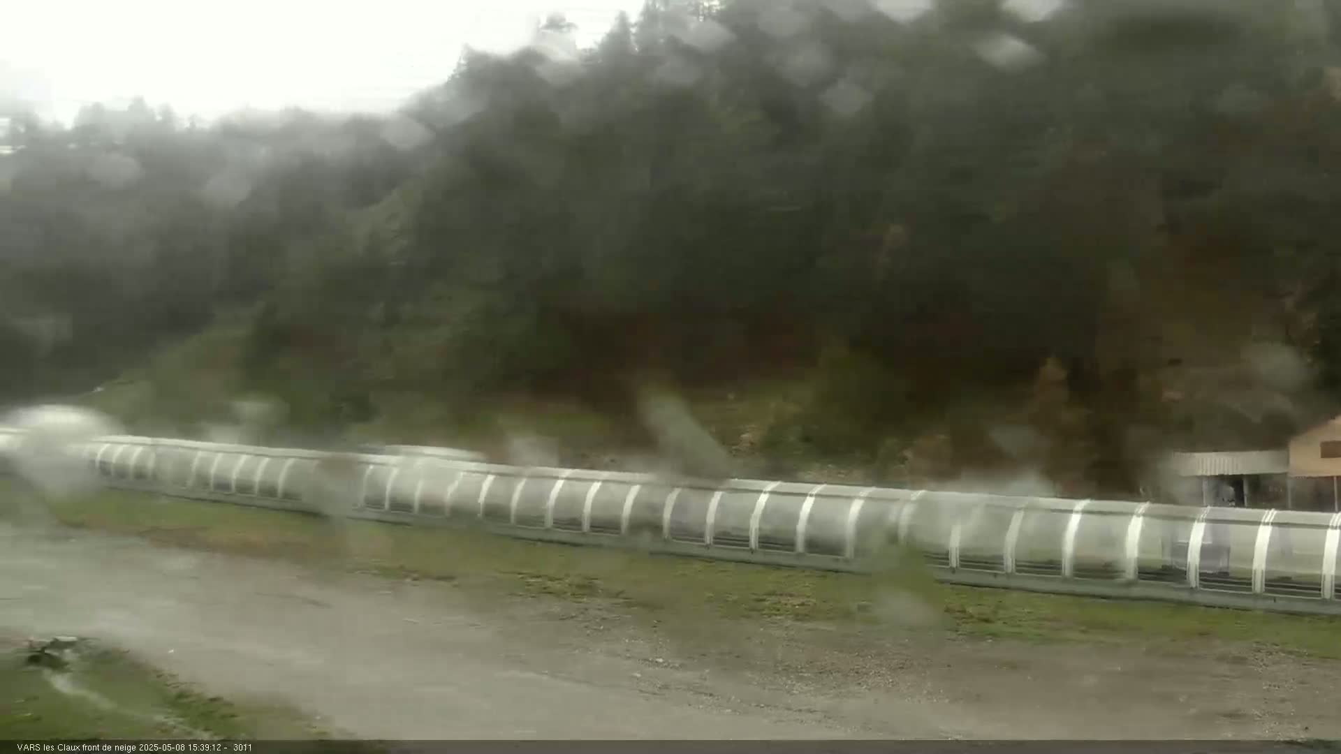A long, covered structure extends across a wet, muddy area, with a wooded hillside visible in the background under a heavy rain.