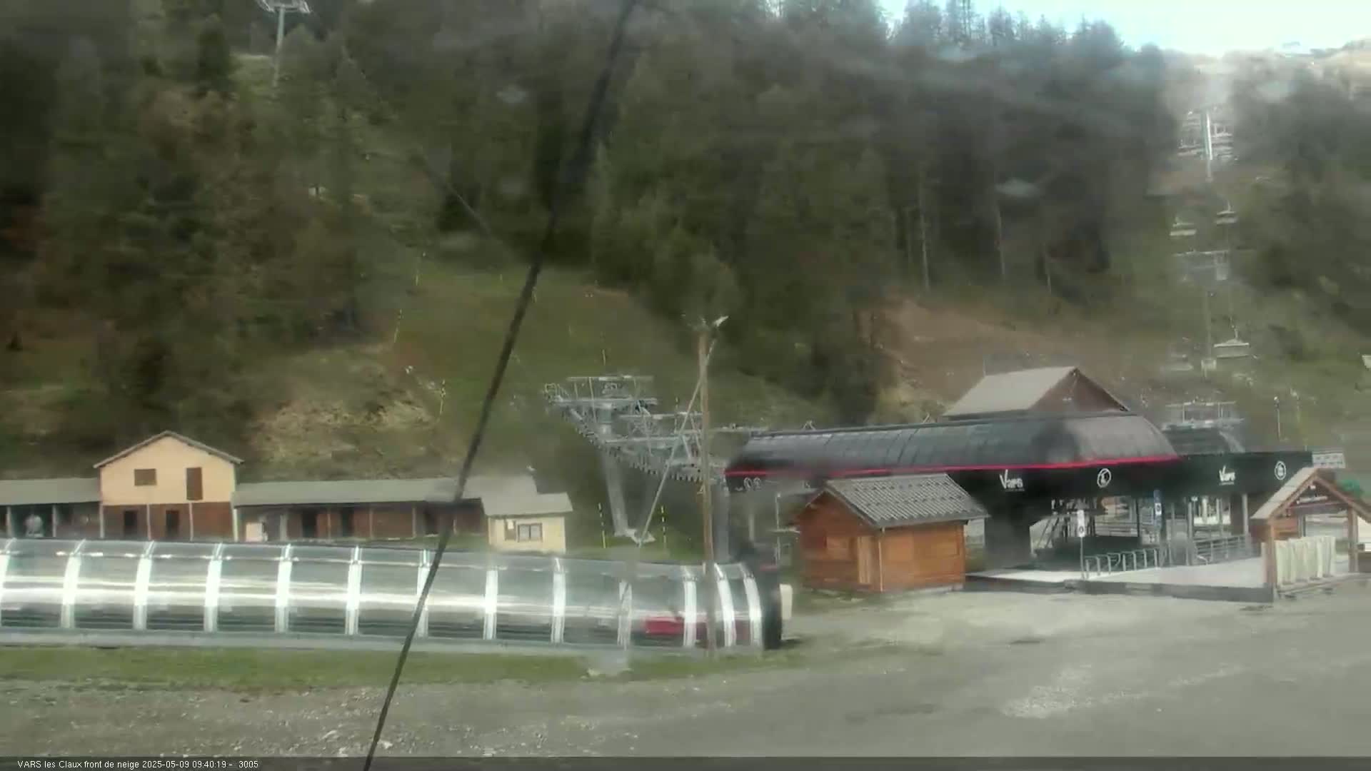 A ski lift base station with several buildings is situated on a hillside covered in trees under overcast skies.
