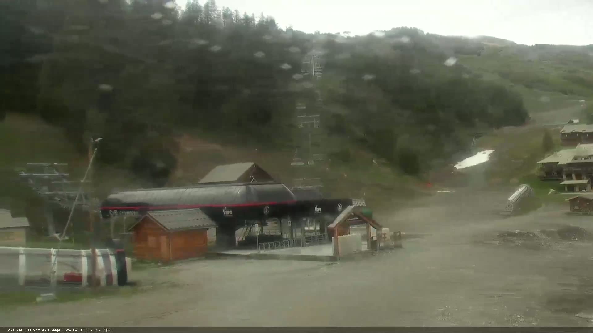 A ski lift base station and surrounding area are visible on a rainy day, with a hillside covered in dark green vegetation in the background.