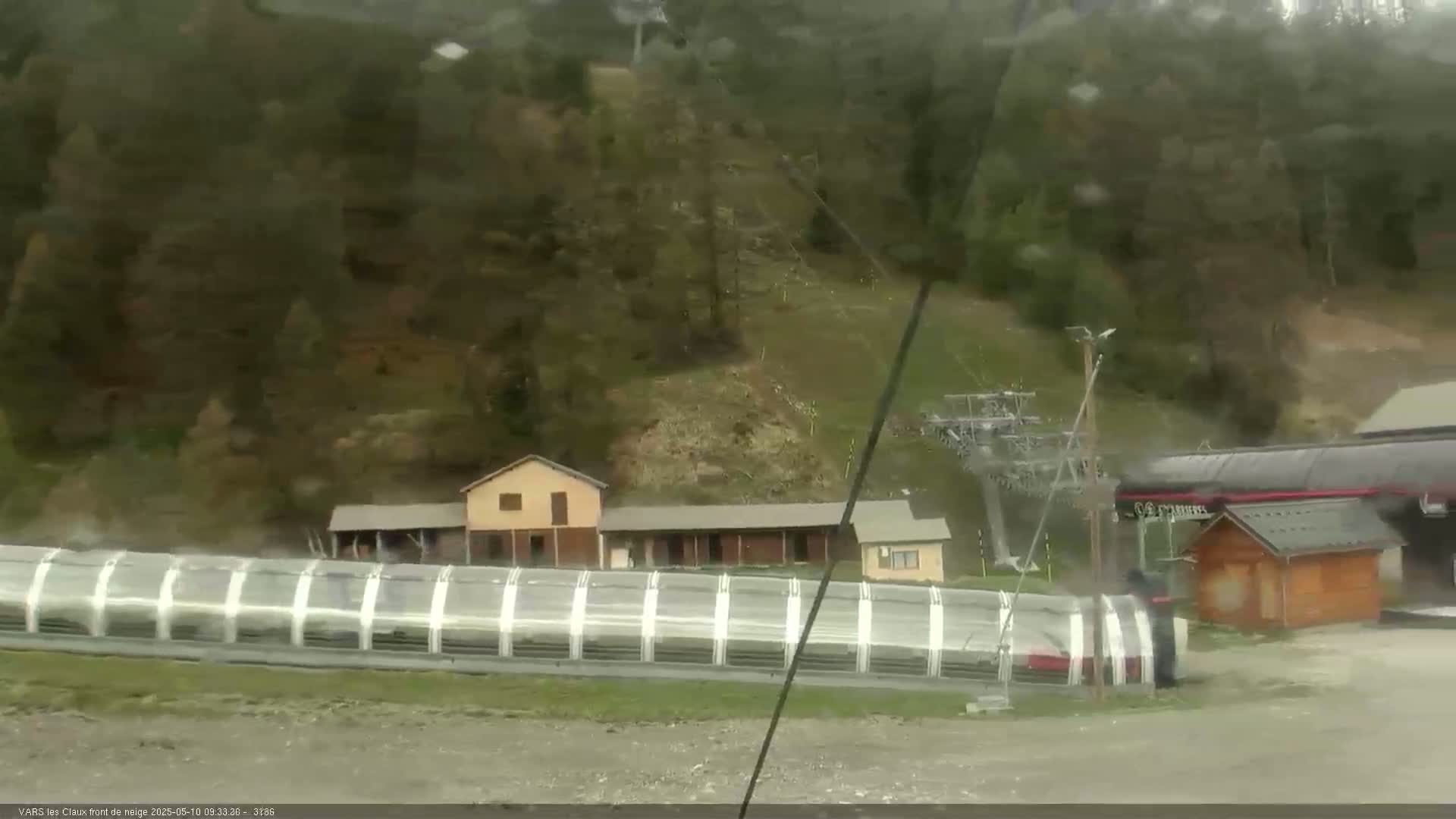 A ski lift and several buildings are visible in the rain, nestled amidst a hillside covered in dark green trees.