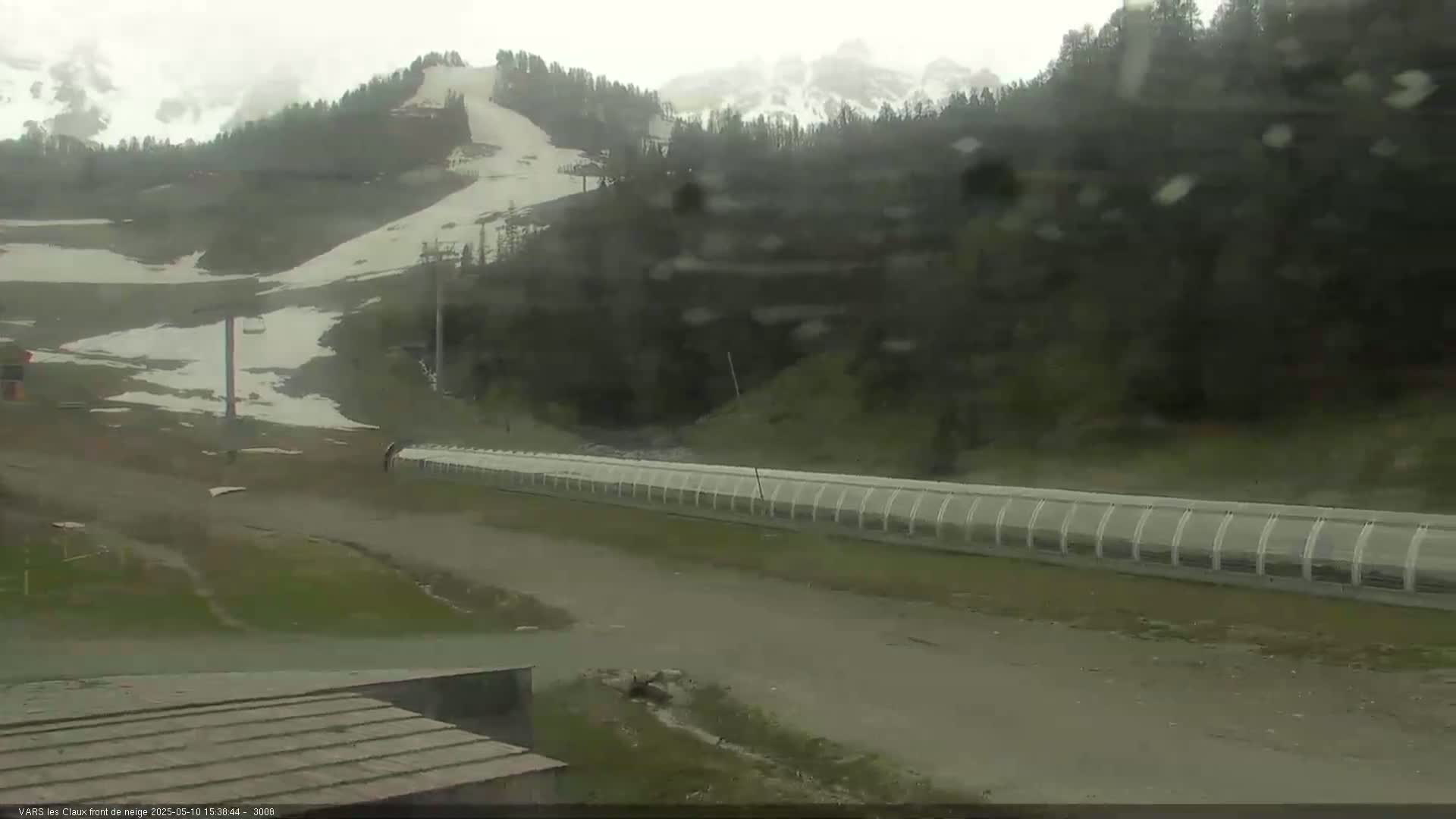A snow-dusted ski slope is visible in the distance, under a rainy overcast sky, with a long protective barrier running across the foreground.