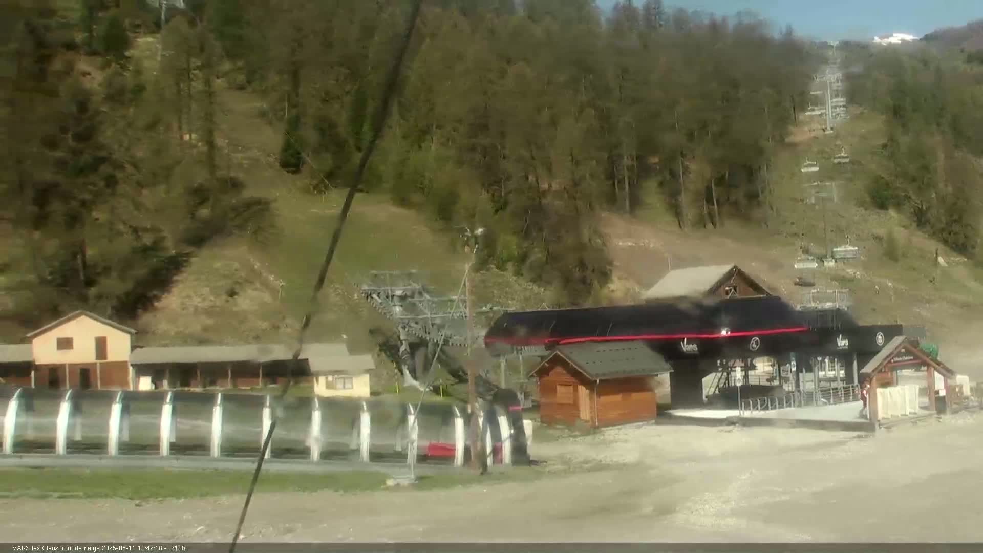 A ski lift base station with several small buildings and a chairlift ascending a hillside covered in trees on a sunny day.