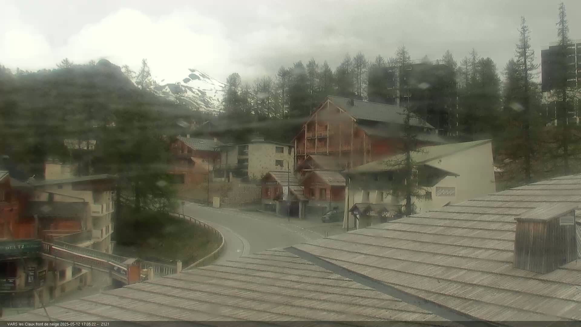 A mountain village is seen on a cloudy day, with snow-capped mountains in the background and rain obscuring the view.