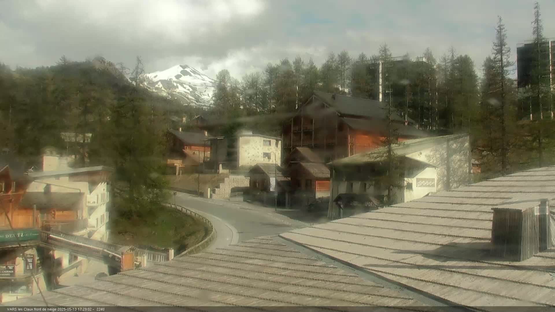 A rain-misted view of a village nestled among mountains, with a snow-capped peak visible in the distance.