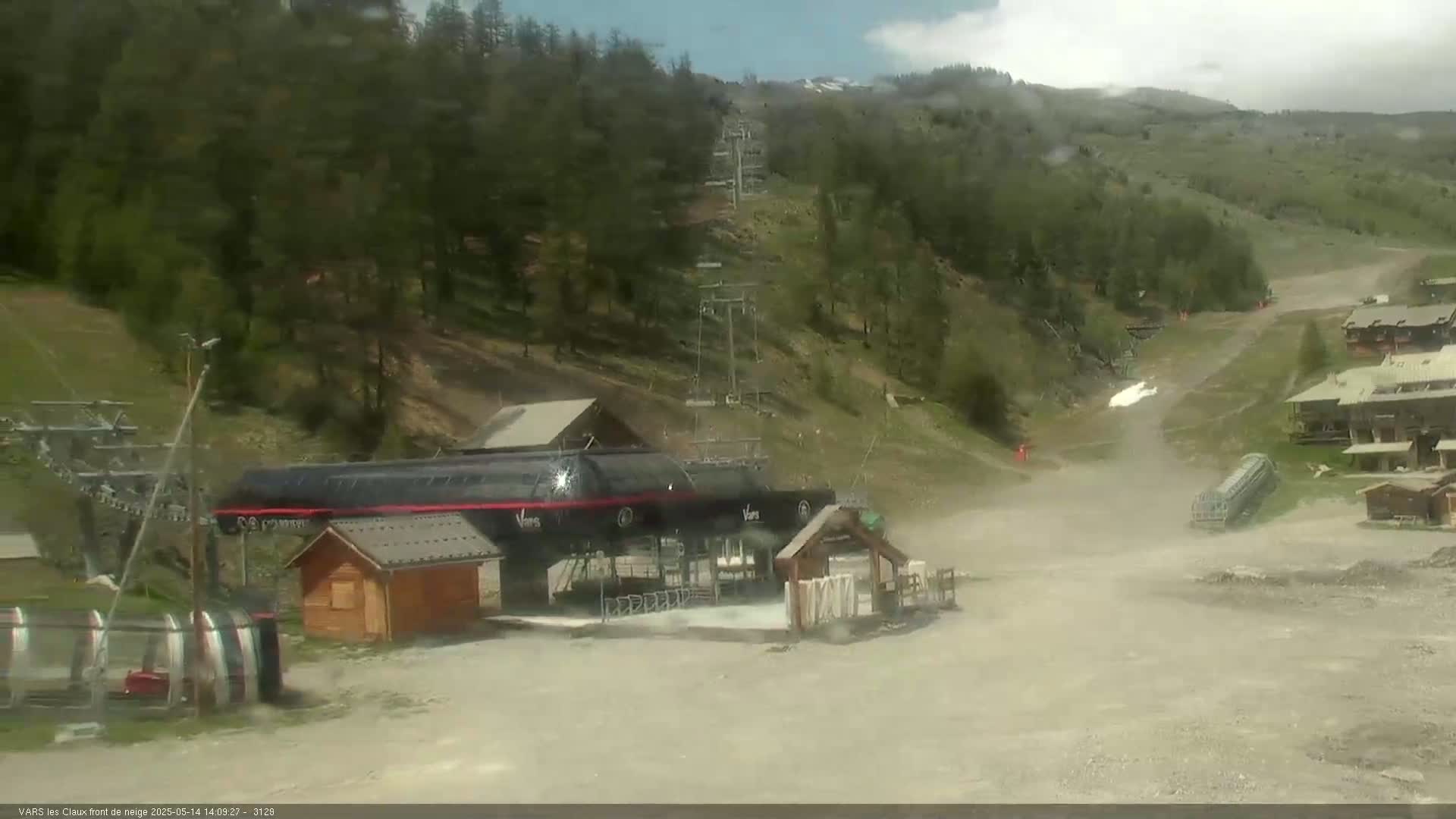 A chairlift base station and surrounding area, with a mostly cloudy sky and some light precipitation, are situated on a hillside covered with trees and patchy, dry ground.