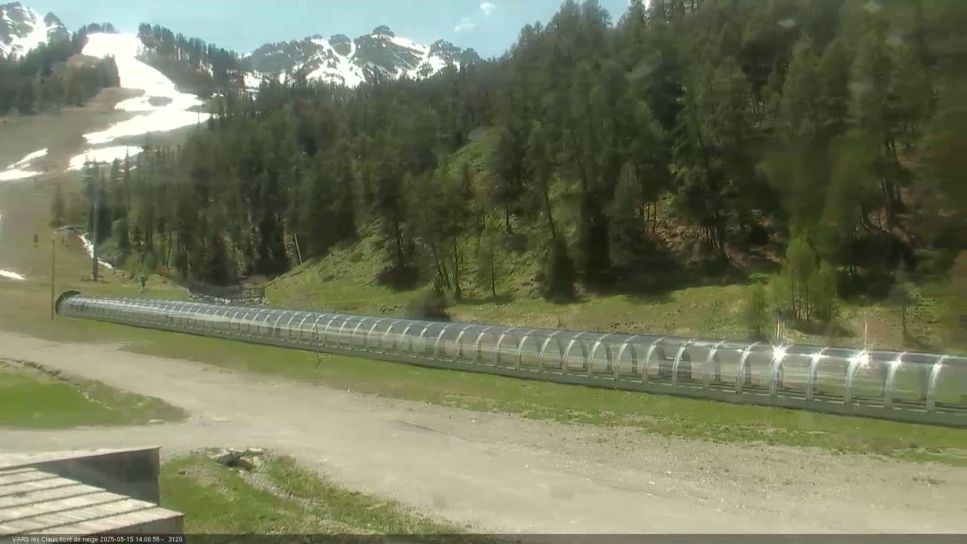 A long, covered conveyor belt sits on a grassy area near a forested hillside with snow-capped mountains in the background under a partly sunny sky.