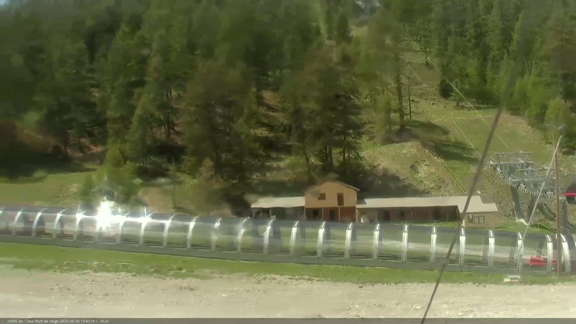 A covered ski lift extends from a building nestled amongst a hillside of evergreen trees on a sunny day.