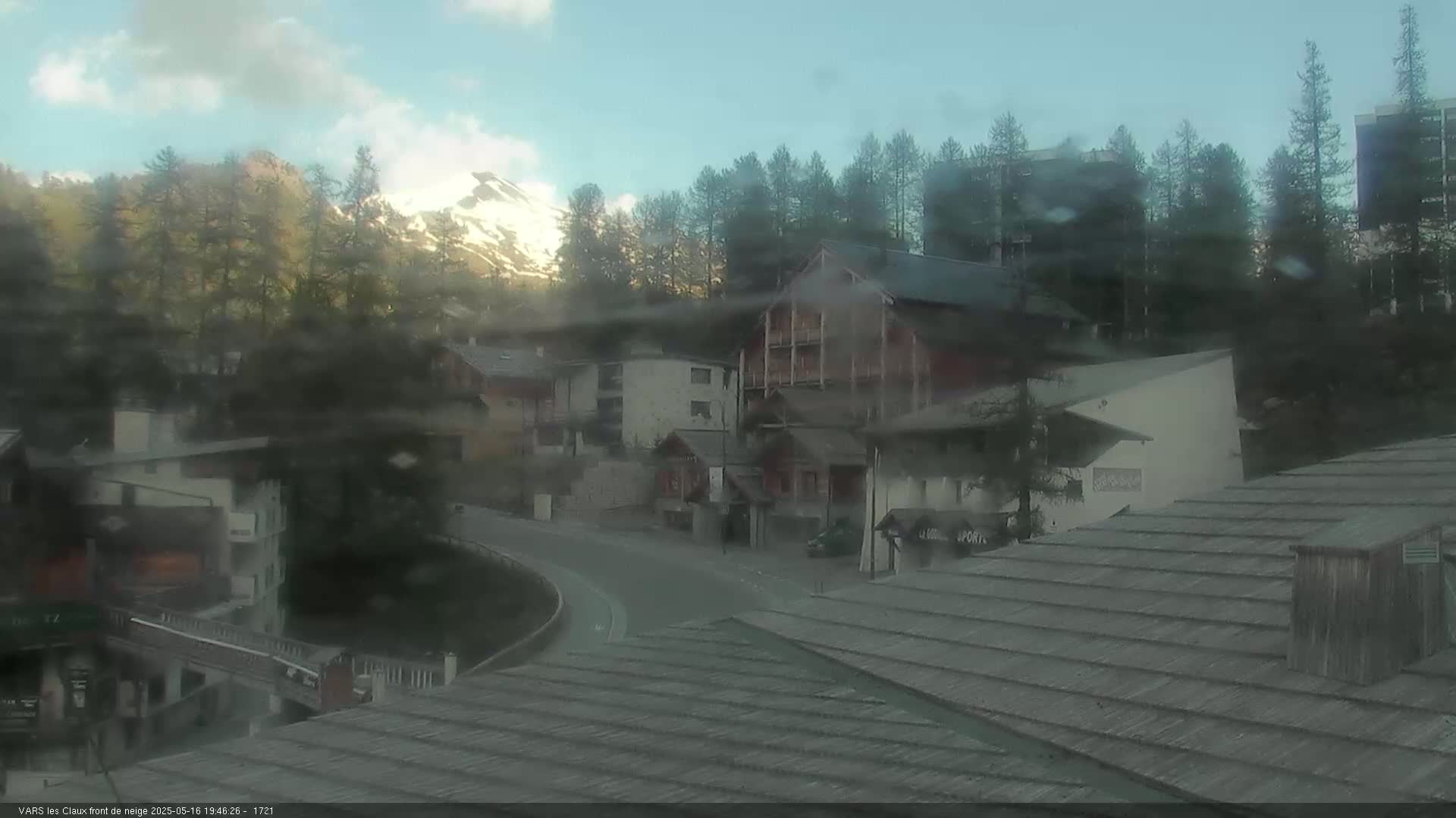 A hazy view of a mountain village with snow-capped mountains in the background under a partly cloudy sky.