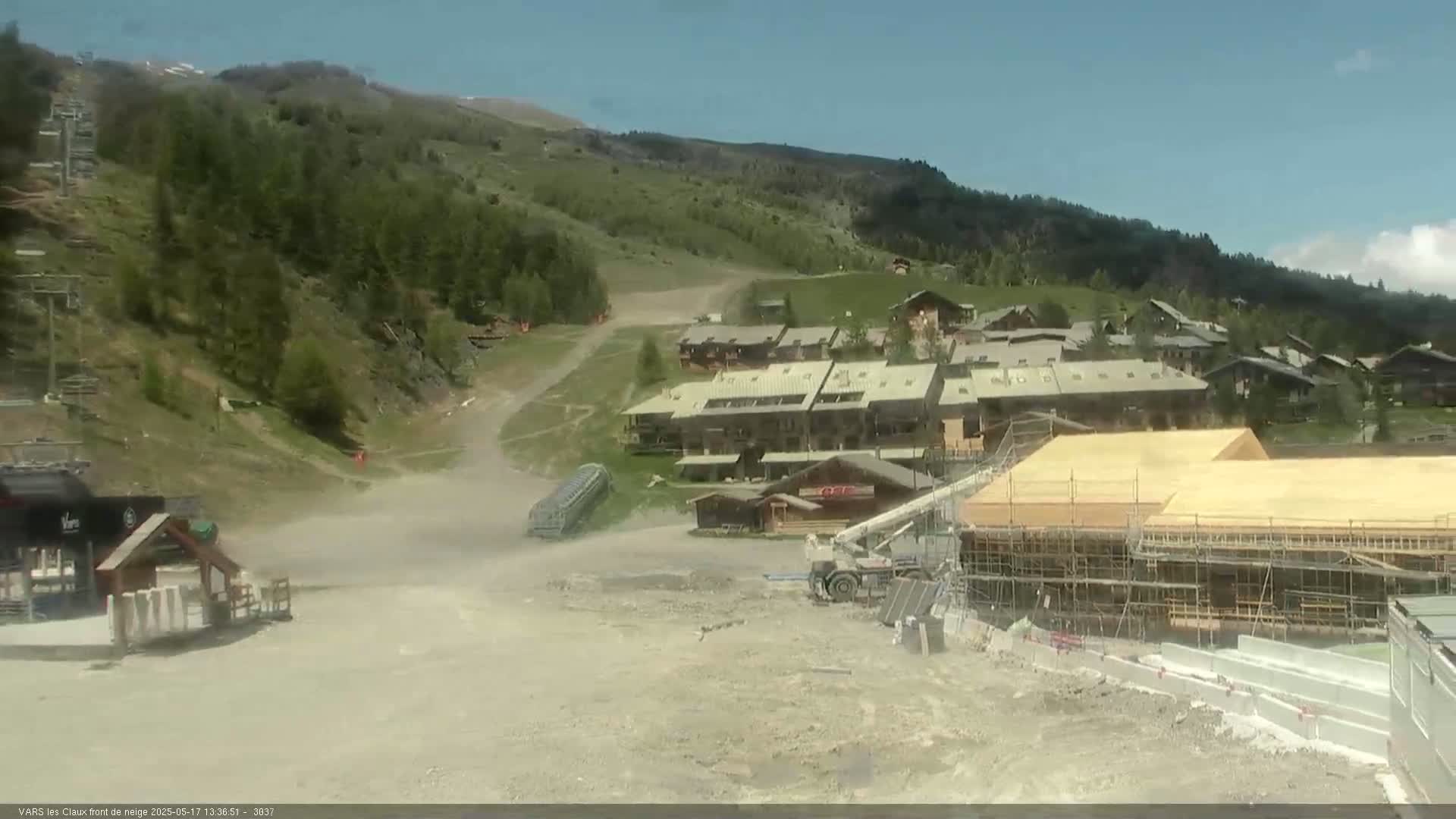 A mountain village with several buildings, including one under construction, is visible on a sunny day, with a dirt road leading up to it and a ski lift in the background.